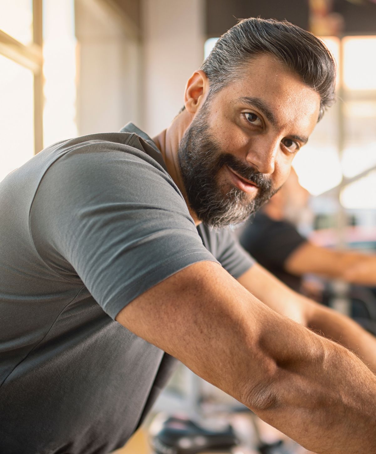 A man with a beard and short hair, wearing a gray shirt, smiling at the camera while resting his arms on a fitness bike.