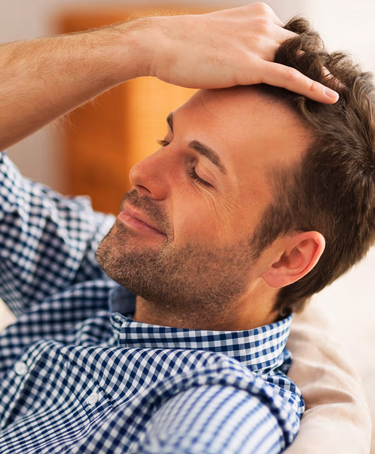 A man with short hair and a beard lightly resting his hand on his forehead while smiling softly, wearing a plaid button-up shirt.
