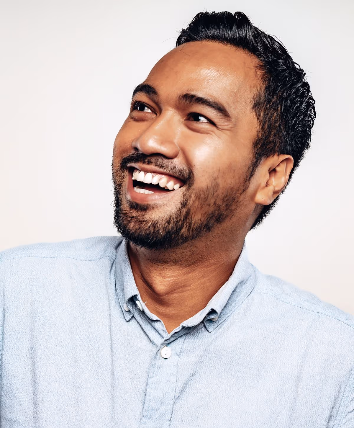 A man with short black hair and a beard smiling broadly while looking slightly upwards. He is wearing a light blue button-down shirt.