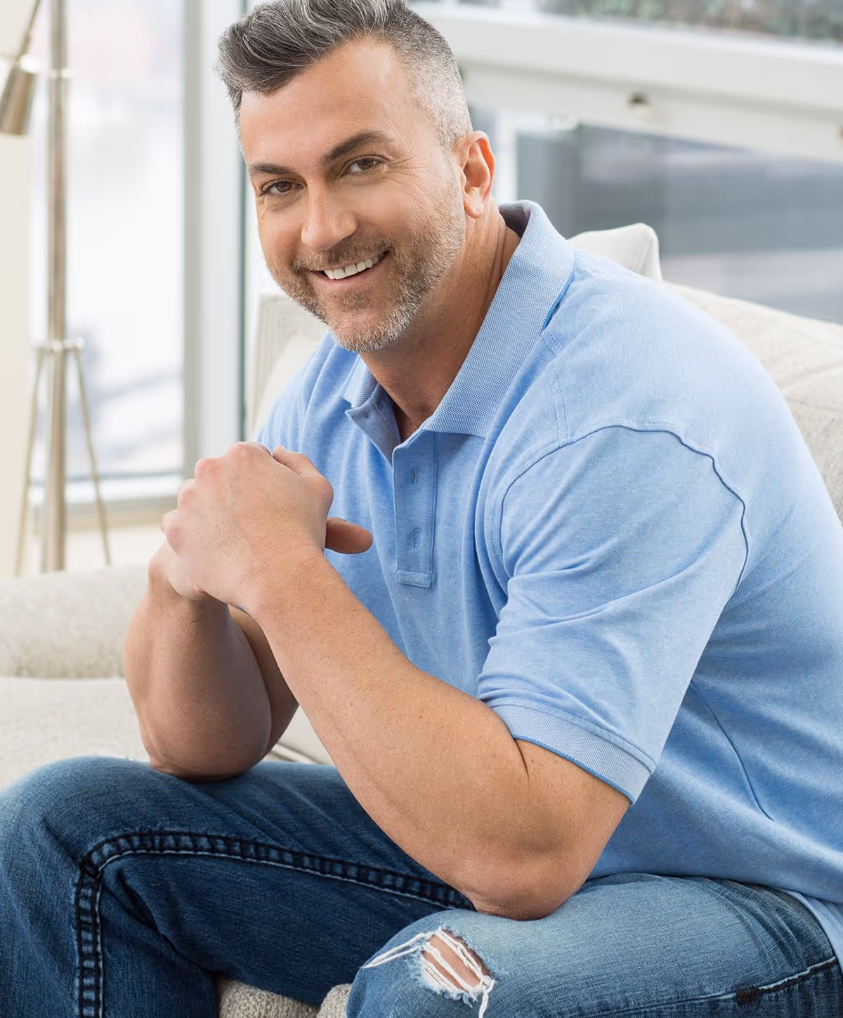 A smiling man with short gray hair, wearing a light blue polo shirt and ripped jeans, sitting on a cream-colored couch.