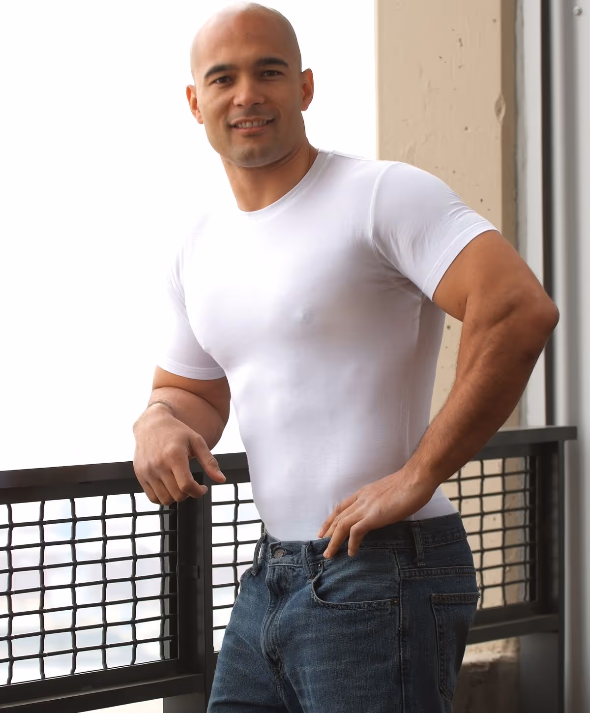 A man wearing a fitted white short-sleeve shirt and blue jeans, standing with one hand on his hip and a confident smile.