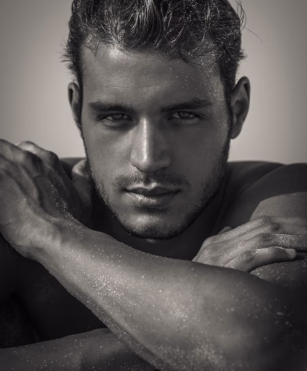 A close-up of a male model with tousled hair, showcasing a serious expression and muscular arms crossed in front of his chest, glistening with water droplets, set against a soft focus background.