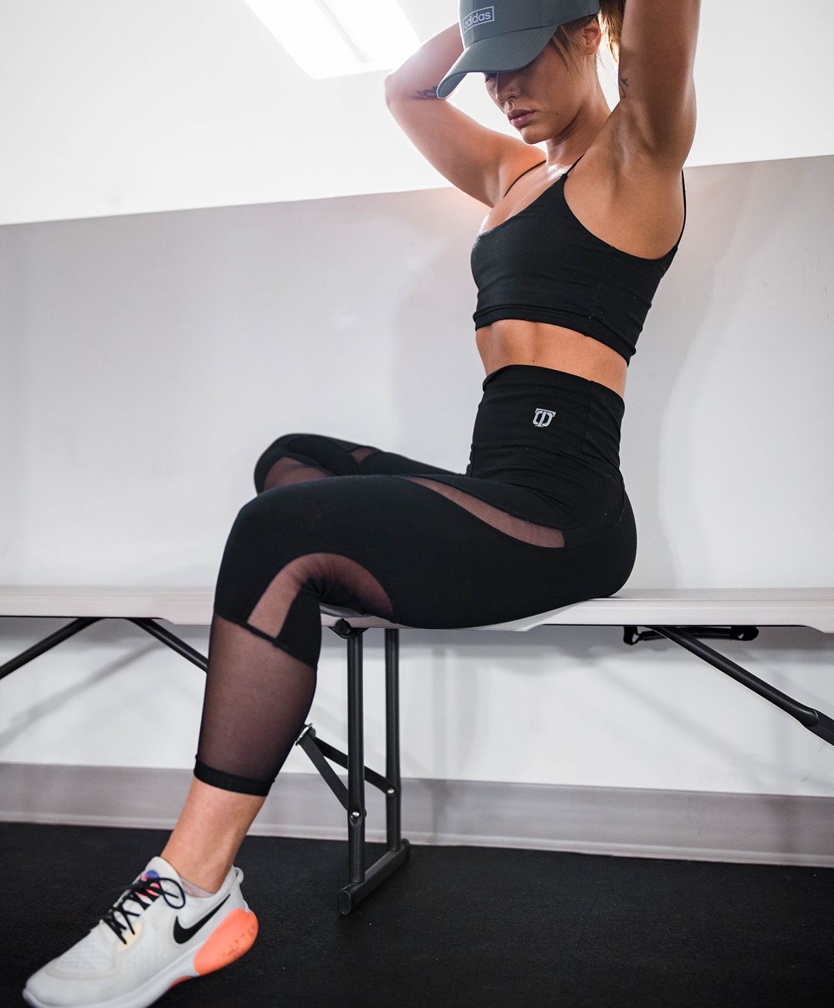 A fit woman sitting on a gym bench wearing a black sports bra and high-waist leggings, with toned abs and arms, in a gym setting with equipment visible in the background.