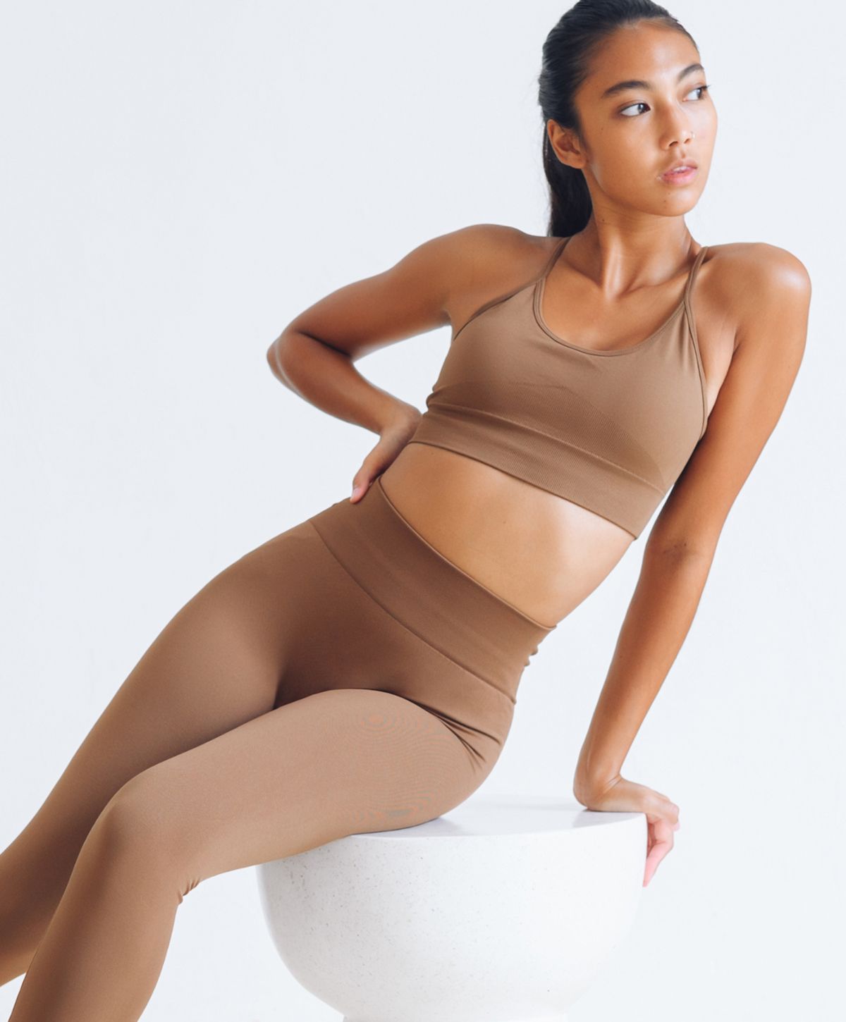 A model wearing a brown sports bra and matching high-waisted leggings, sitting on a round white pedestal, with a relaxed pose.