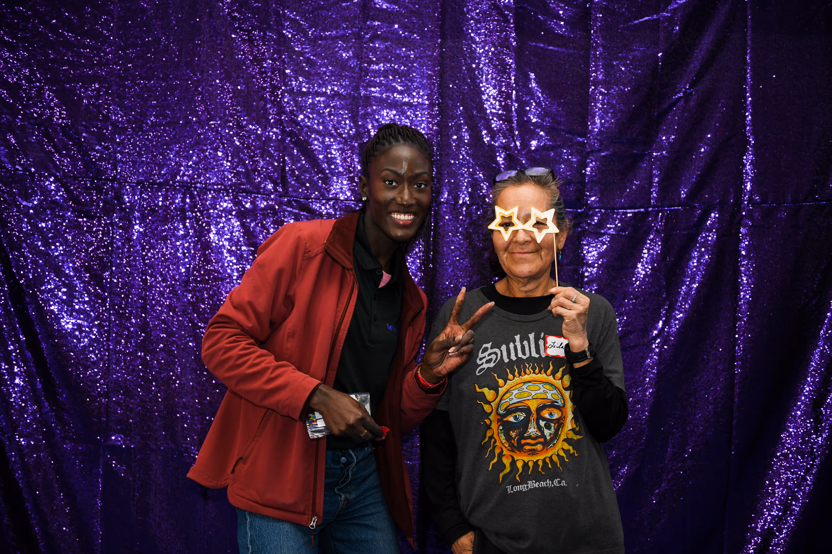 Two women posing against a shiny purple backdrop, one wearing a red jacket and smiling, the other wearing star-shaped glasses and a Sublime band t-shirt.