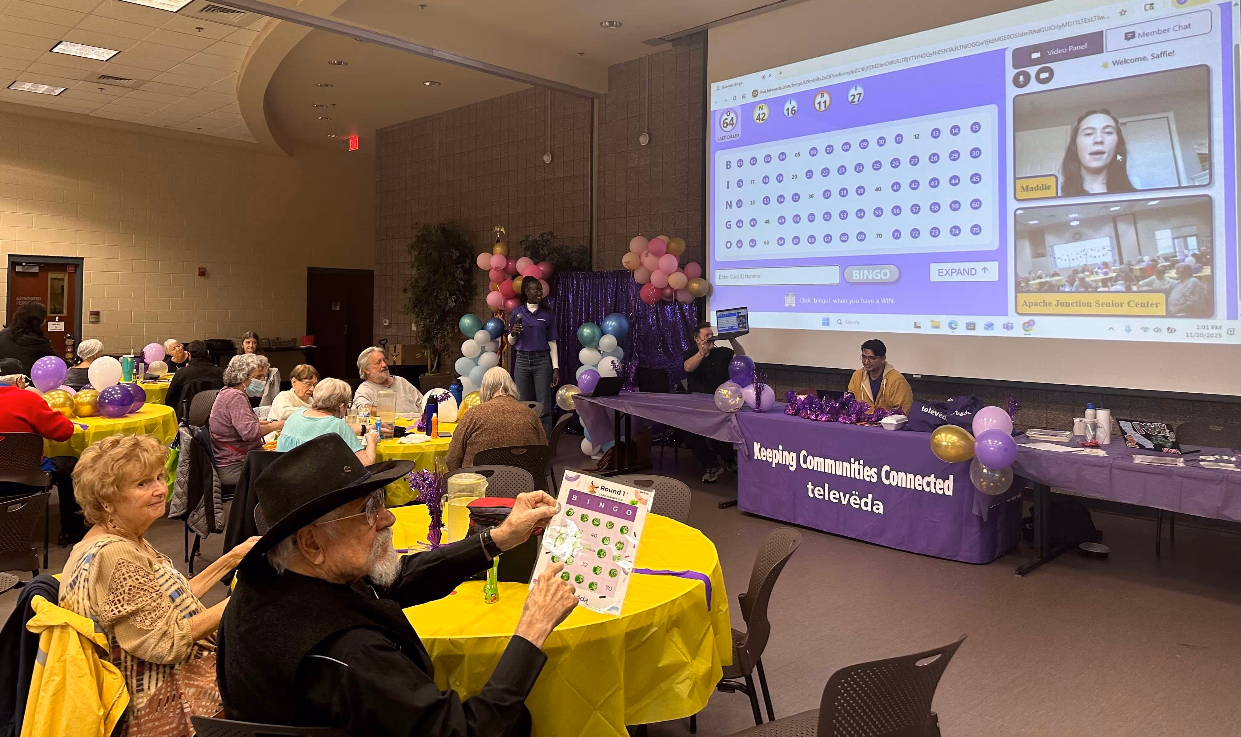 Elderly people playing virtual bingo in a community center with a large screen showing a bingo card and video chat.