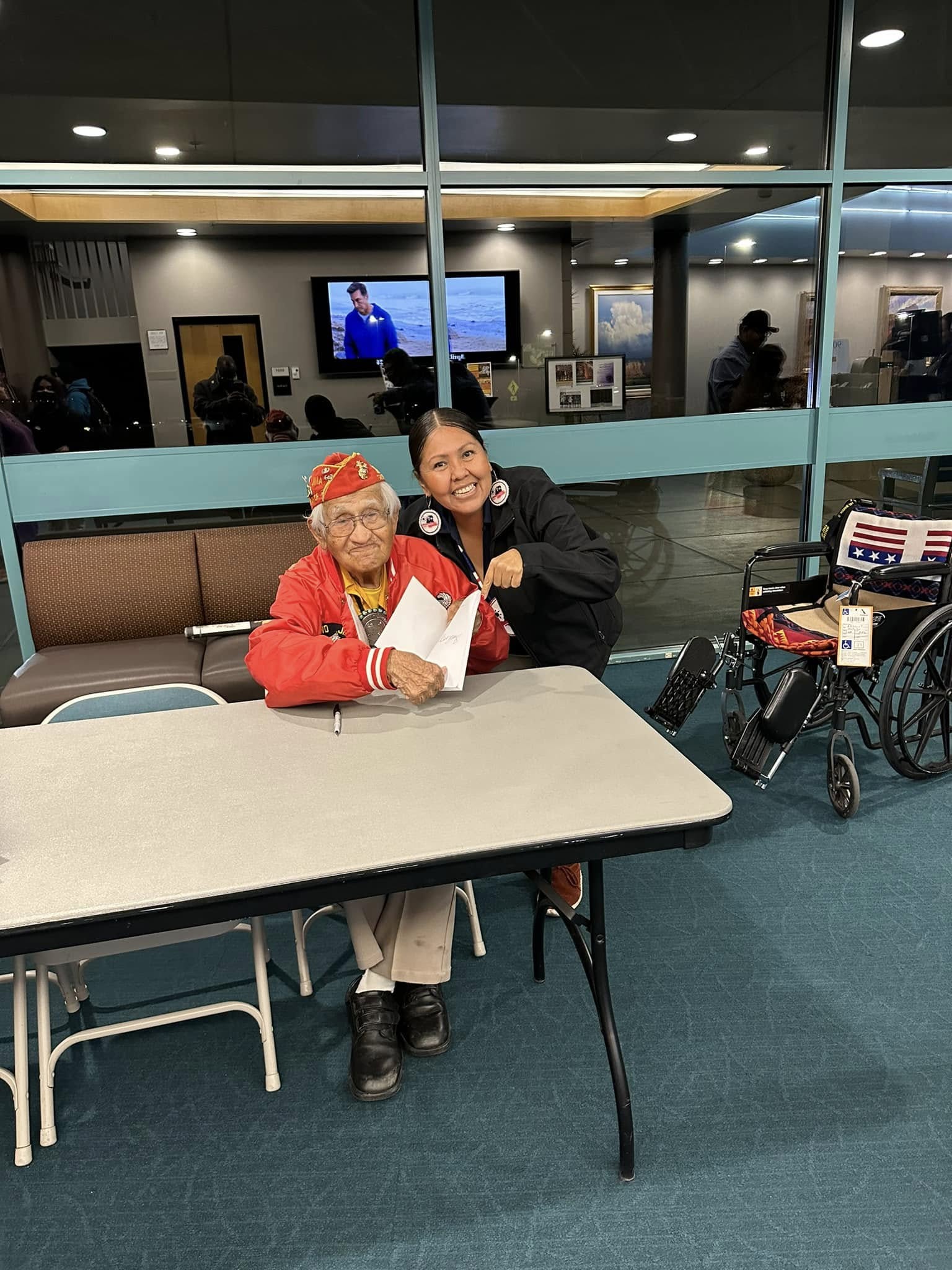 Older woman in red and a woman seated at a table in a community room