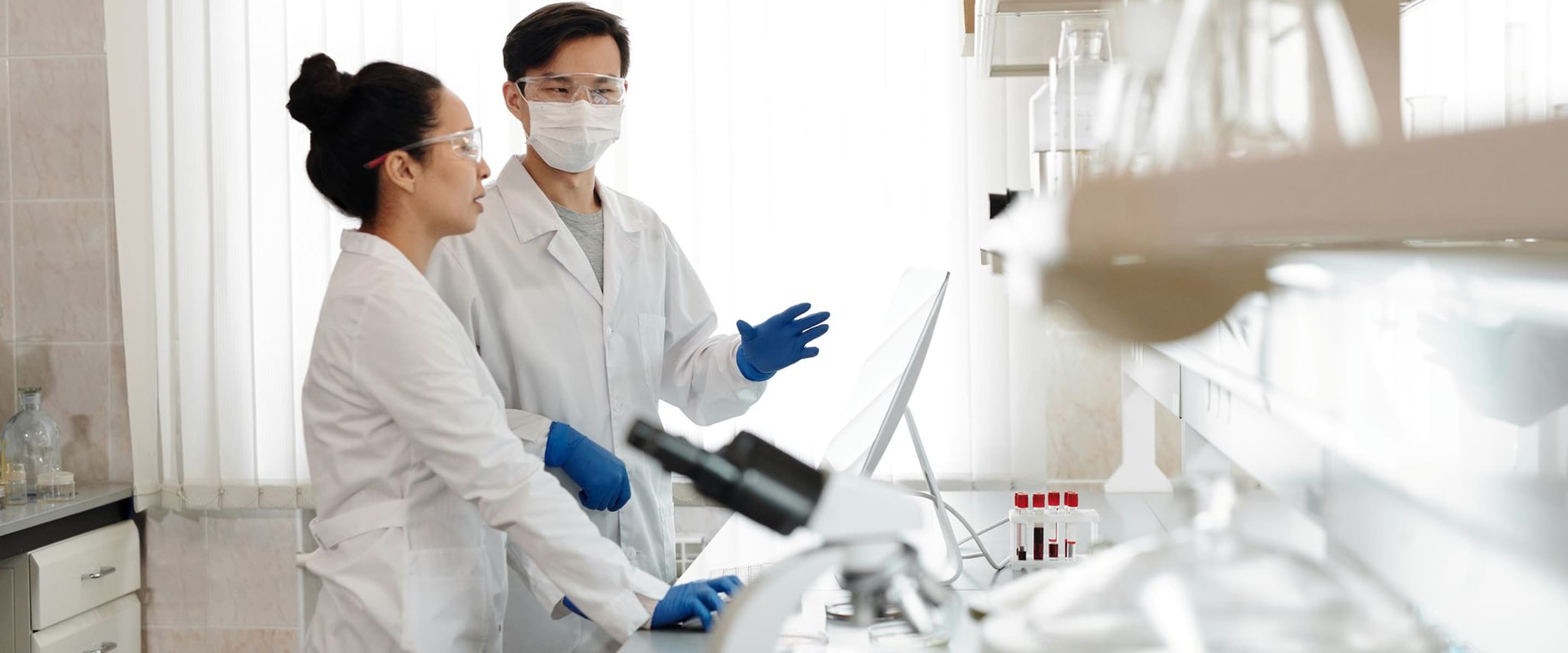 Dva vědci v laboratoři s bílými plášti, rukavicemi a brýlemi diskutují u počítače.