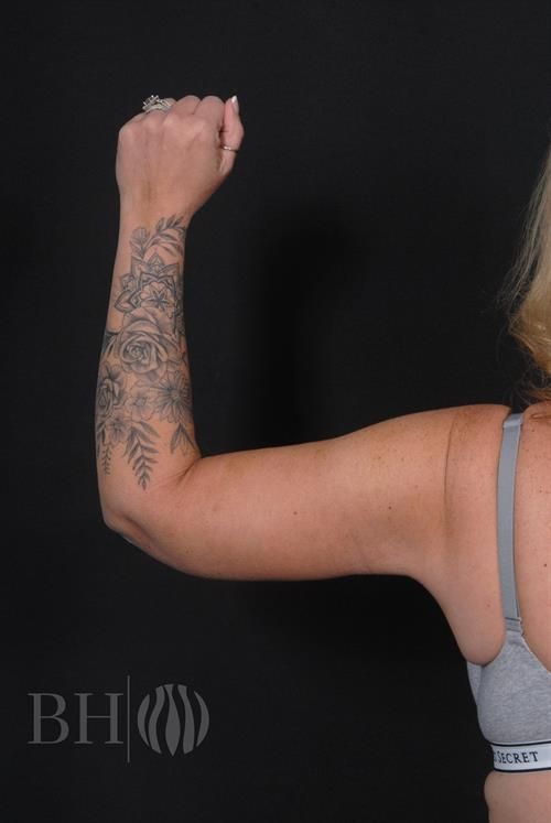 A woman flexing her arm to show her bicep, wearing a gray sports bra. Her forearm features a detailed black and gray floral tattoo with roses, mandalas, and botanical elements. She is wearing rings on her fingers. A watermark logo reading 'BH' with a wave