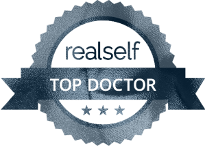 A circular badge with the text 'realself' at the top and 'TOP DOCTOR' below it, accompanied by three stars at the bottom.
