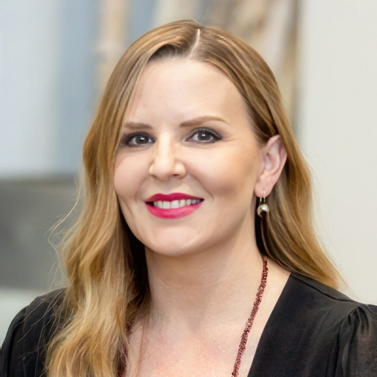 A portrait of a woman with long, wavy blonde hair, wearing a black top and a red necklace, smiling at the camera.