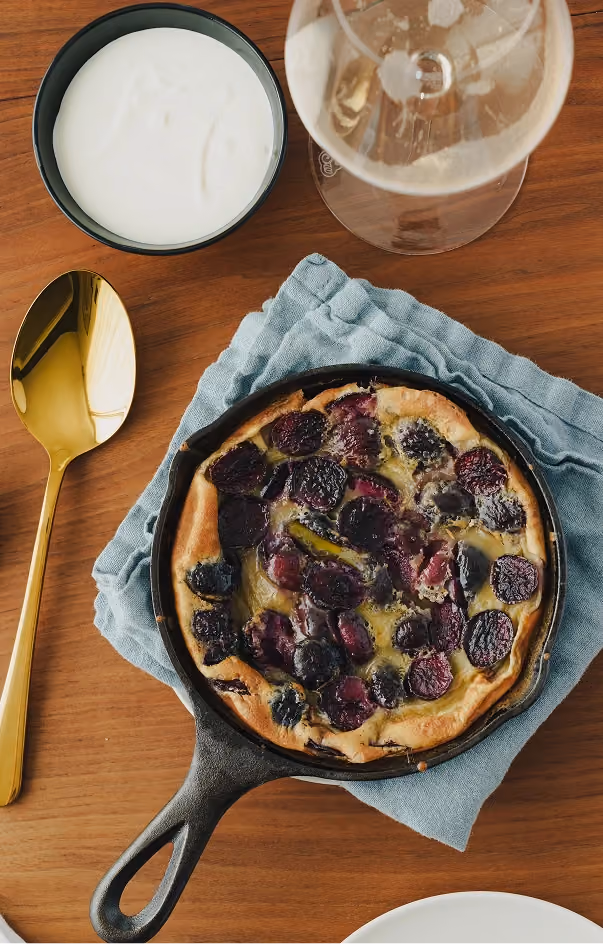 Dark fruit clafoutis in a cast-iron skillet with cream and a glass of Champagne