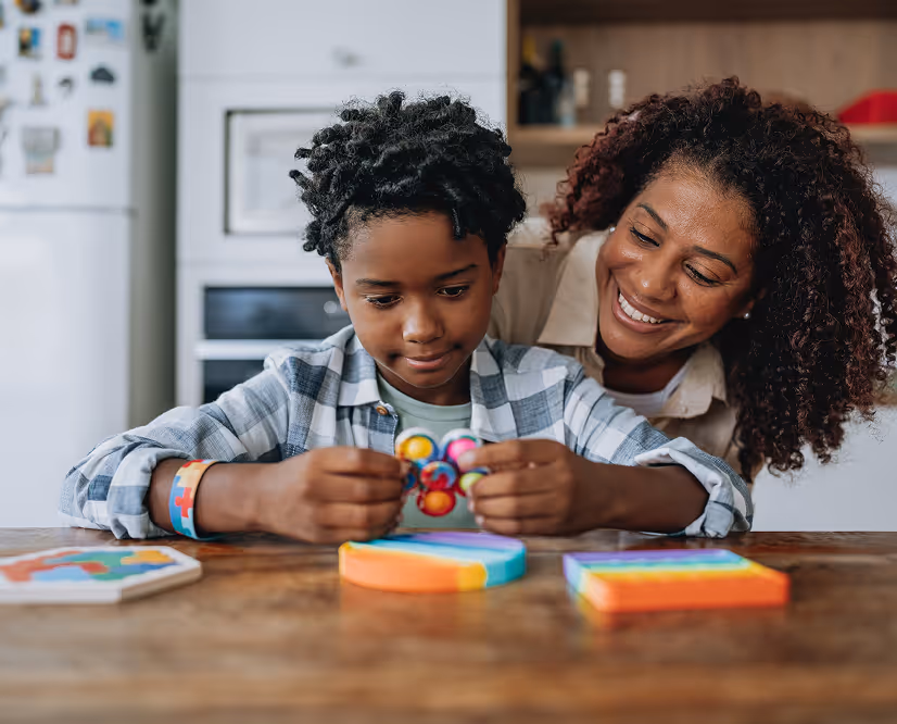 Smiling woman watches young boy holding colorful magnets at a wooden table in a kitchen.