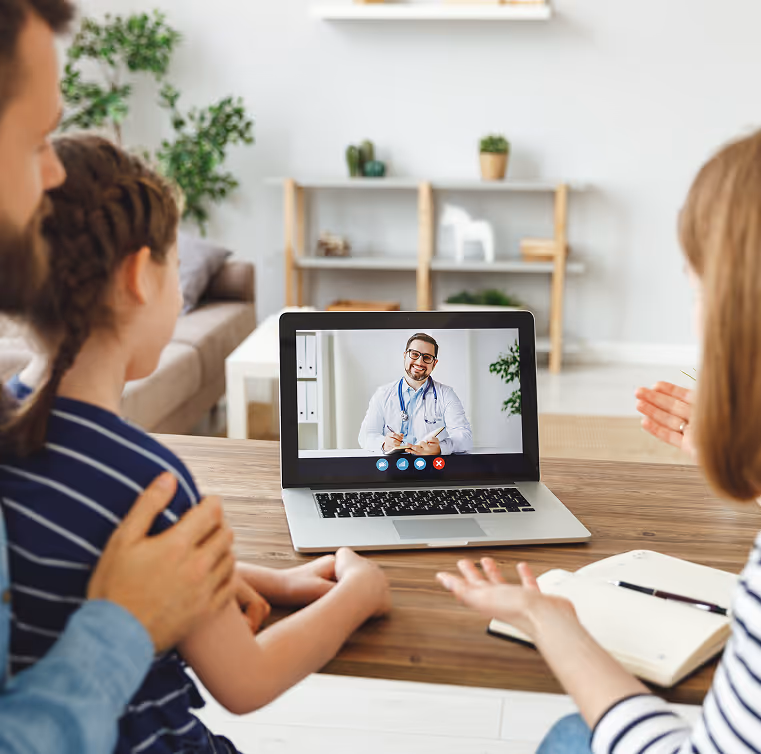 Family video calling a smiling doctor on a laptop for a virtual medical consultation.