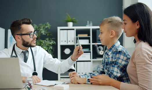 Doctor holding a medical tool while examining a young boy seated next to a woman.