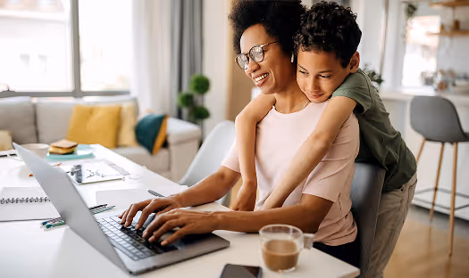 Smiling woman typing on a laptop as a boy hugs her from behind in a bright living room.