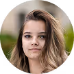 Young woman with long light brown hair and neutral expression in a blurred outdoor setting.