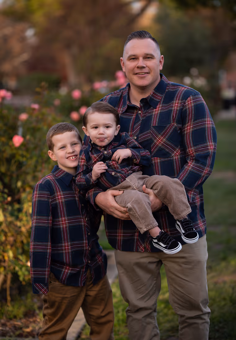 Man holding toddler and standing next to young boy, all in matching plaid shirts in a garden with flowers.