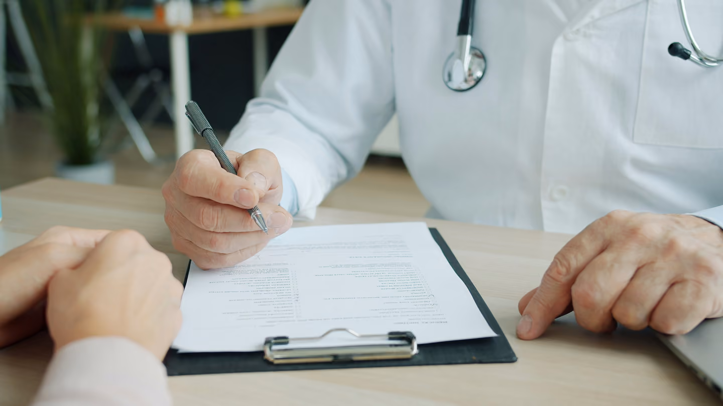 Doctor in white coat holding pen, discussing and pointing to a document on a clipboard with a patient.