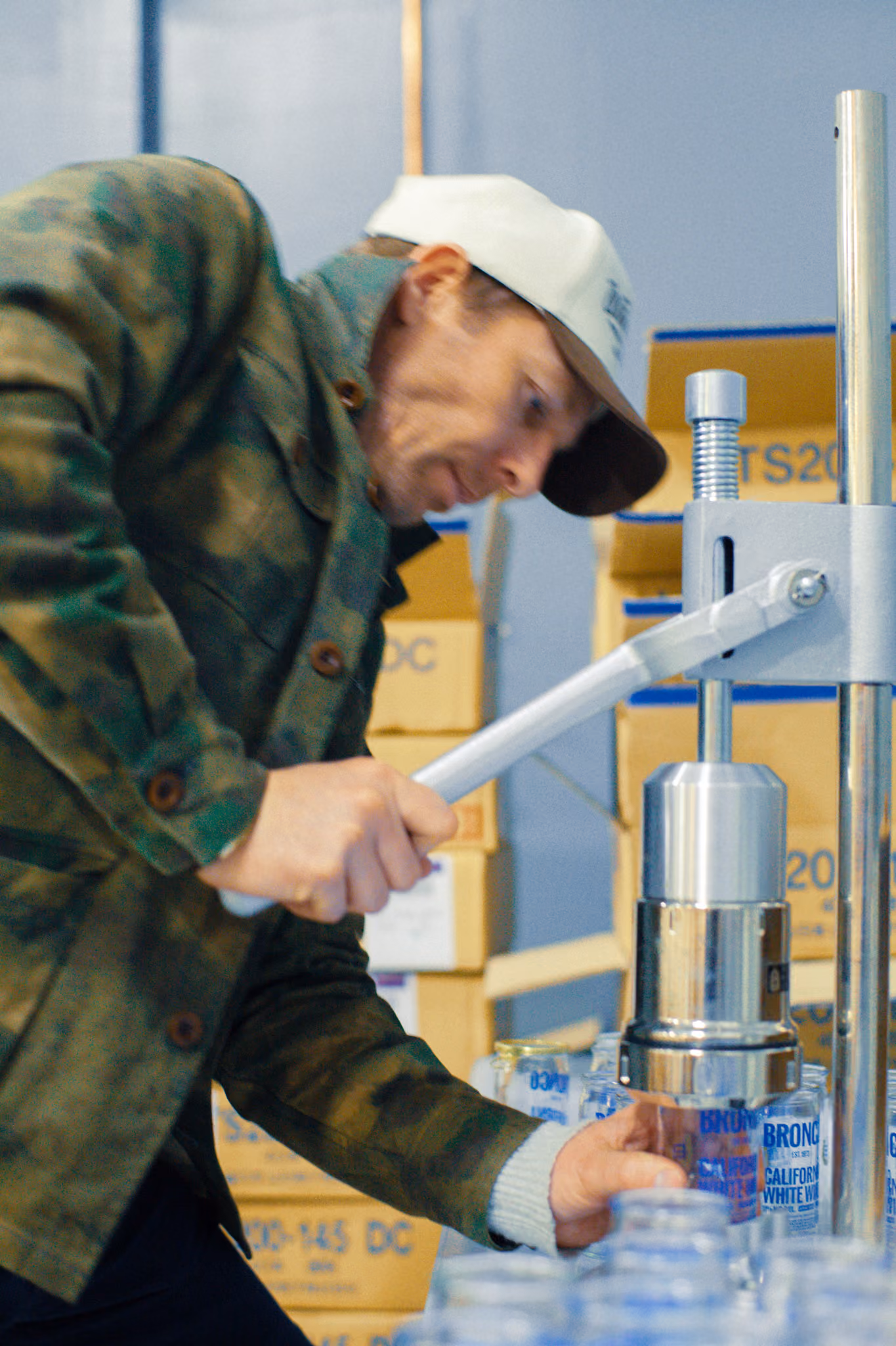 Man in camo jacket and cap using a press to seal lids on clear jars in a warehouse.