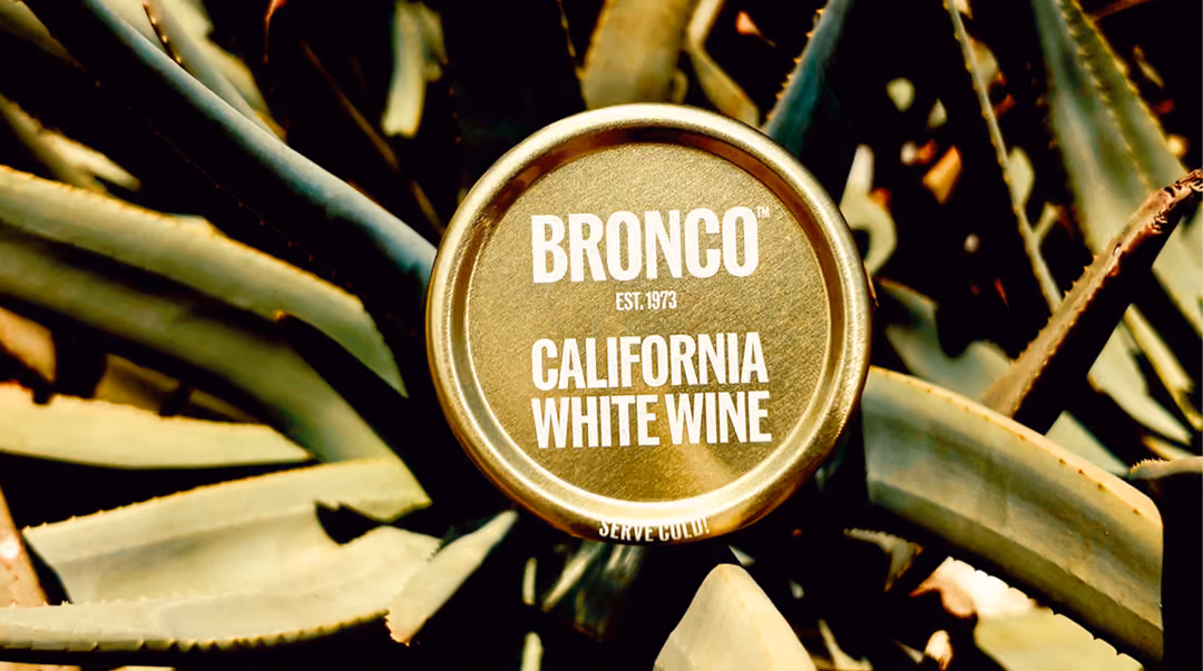Top view of a gold wine bottle cap labeled Bronco California White Wine est. 1973 among green leaves.