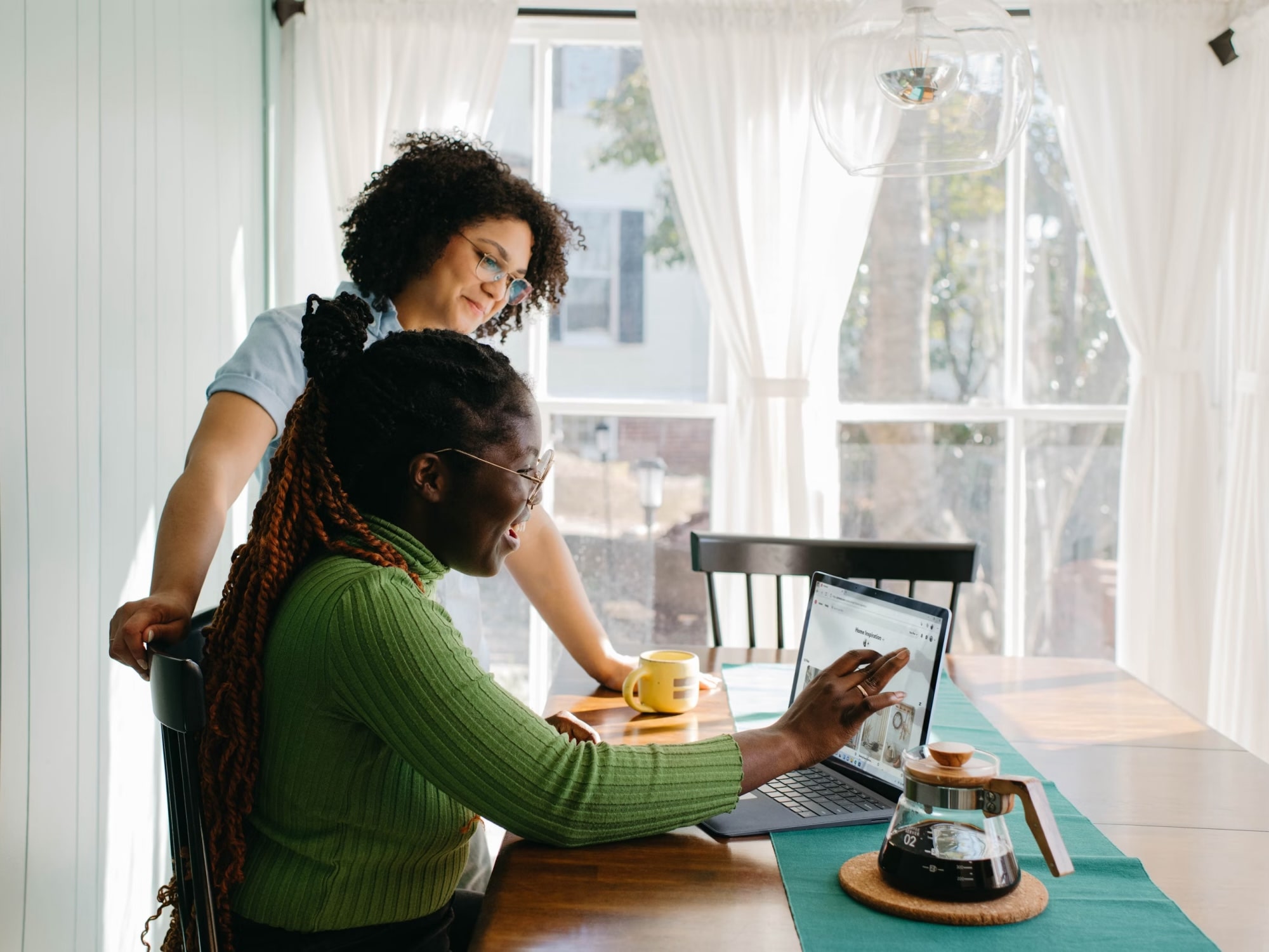 Two women using a laptop together at a sunlit table with a coffee pot and cup nearby.