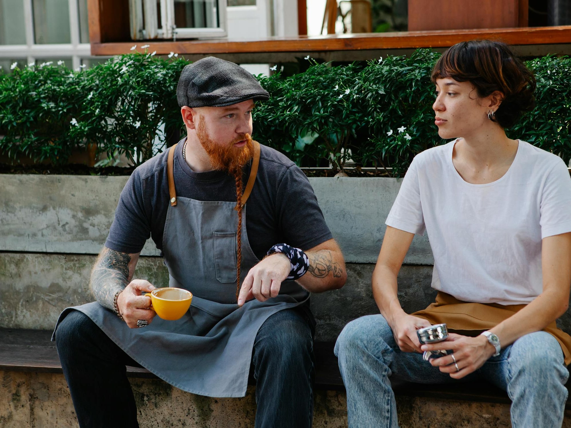 A man in a gray apron and cap holding a yellow cup talks to a woman in a white shirt and jeans.