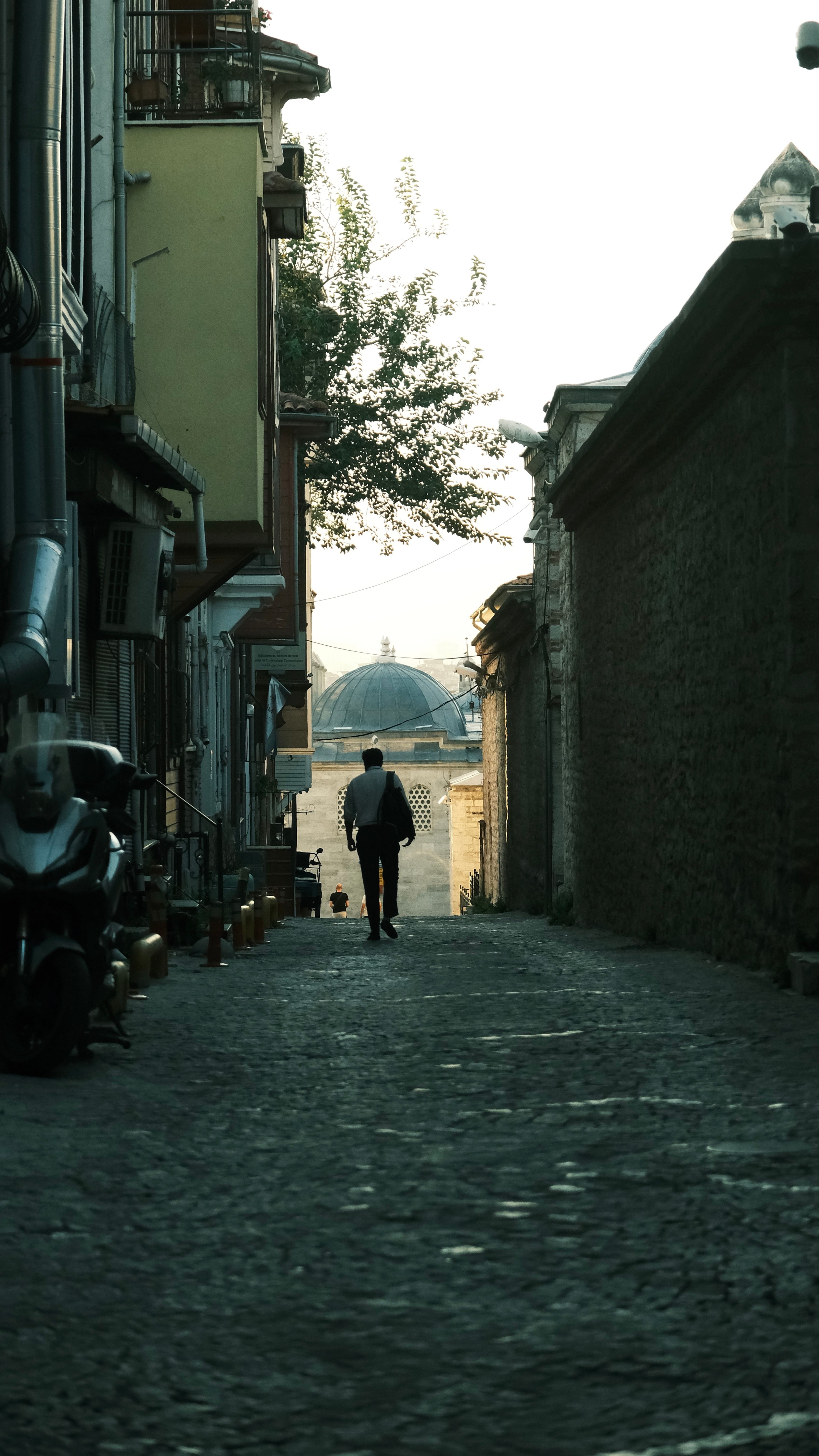 Person walking down a narrow cobblestone street lined with buildings and a dome visible ahead.