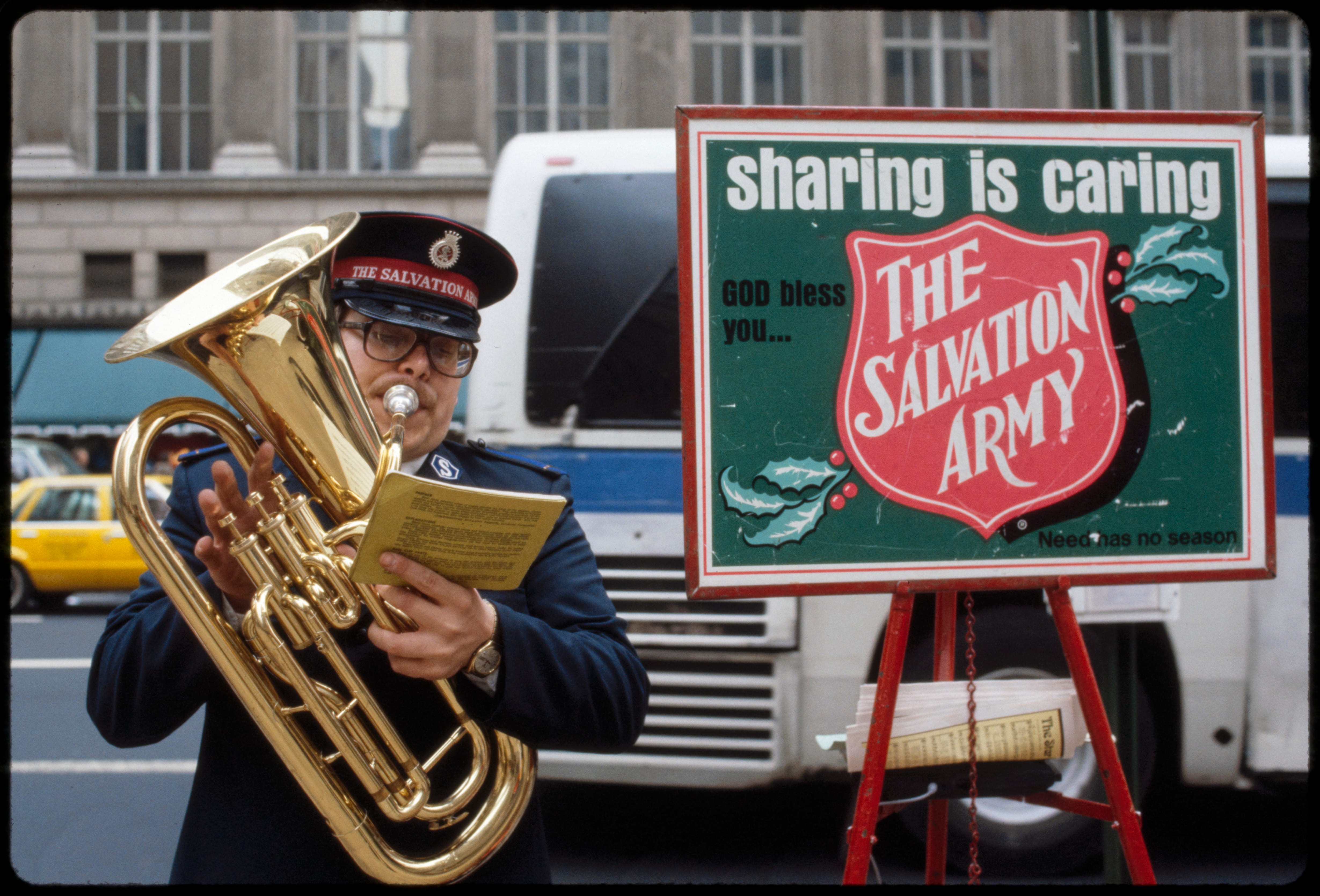 Salvation Army member playing a brass instrument next to a sign reading 'sharing is caring'.