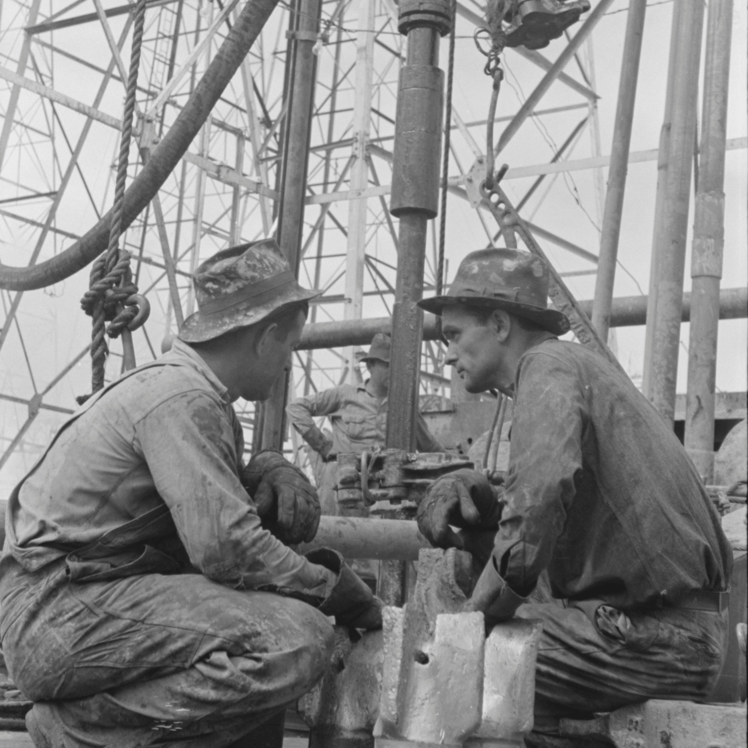 Two workers in hats and dirty clothes crouch near heavy machinery on an industrial site.