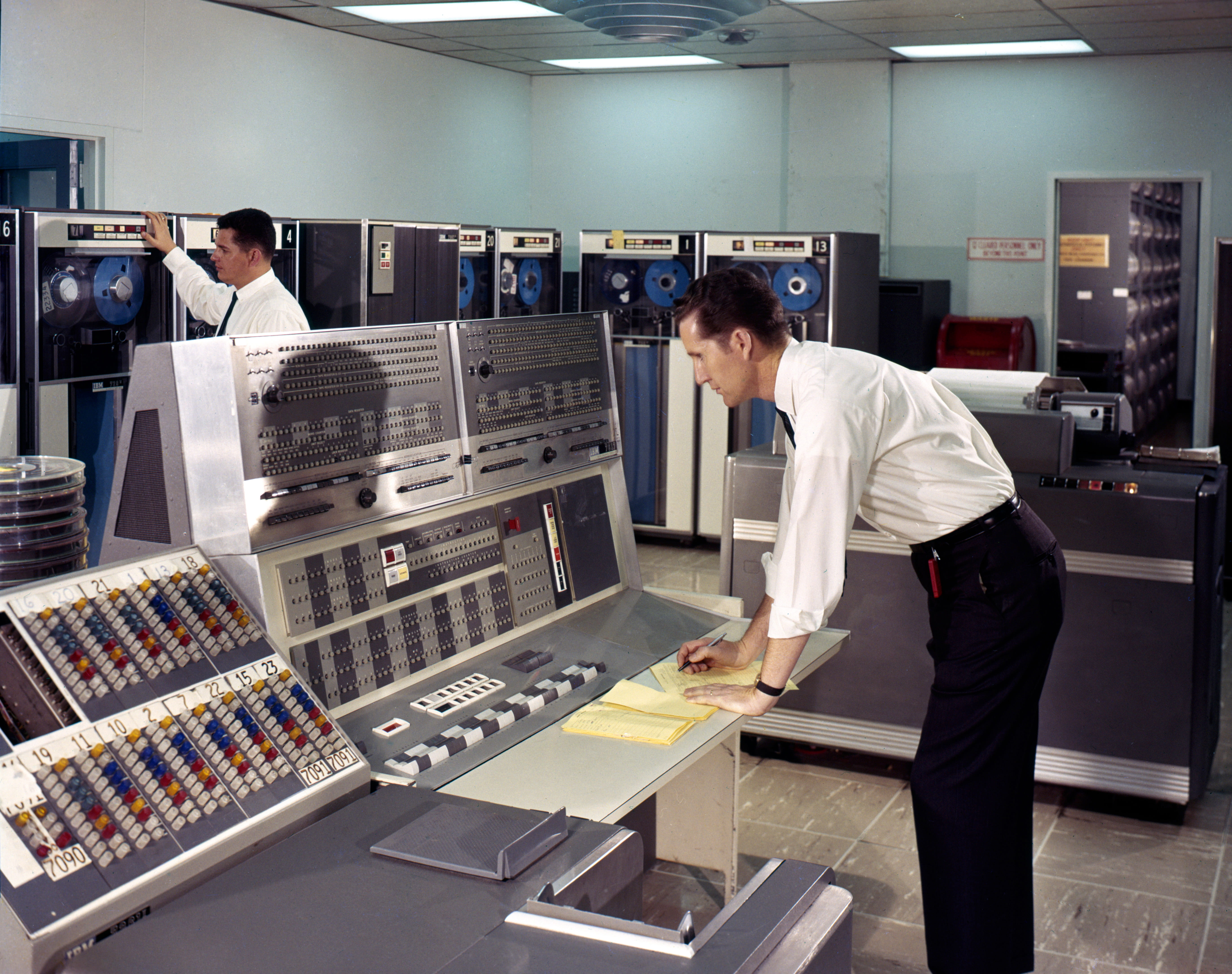 Two men working with vintage mainframe computers with tape drives and control panels in a data center.