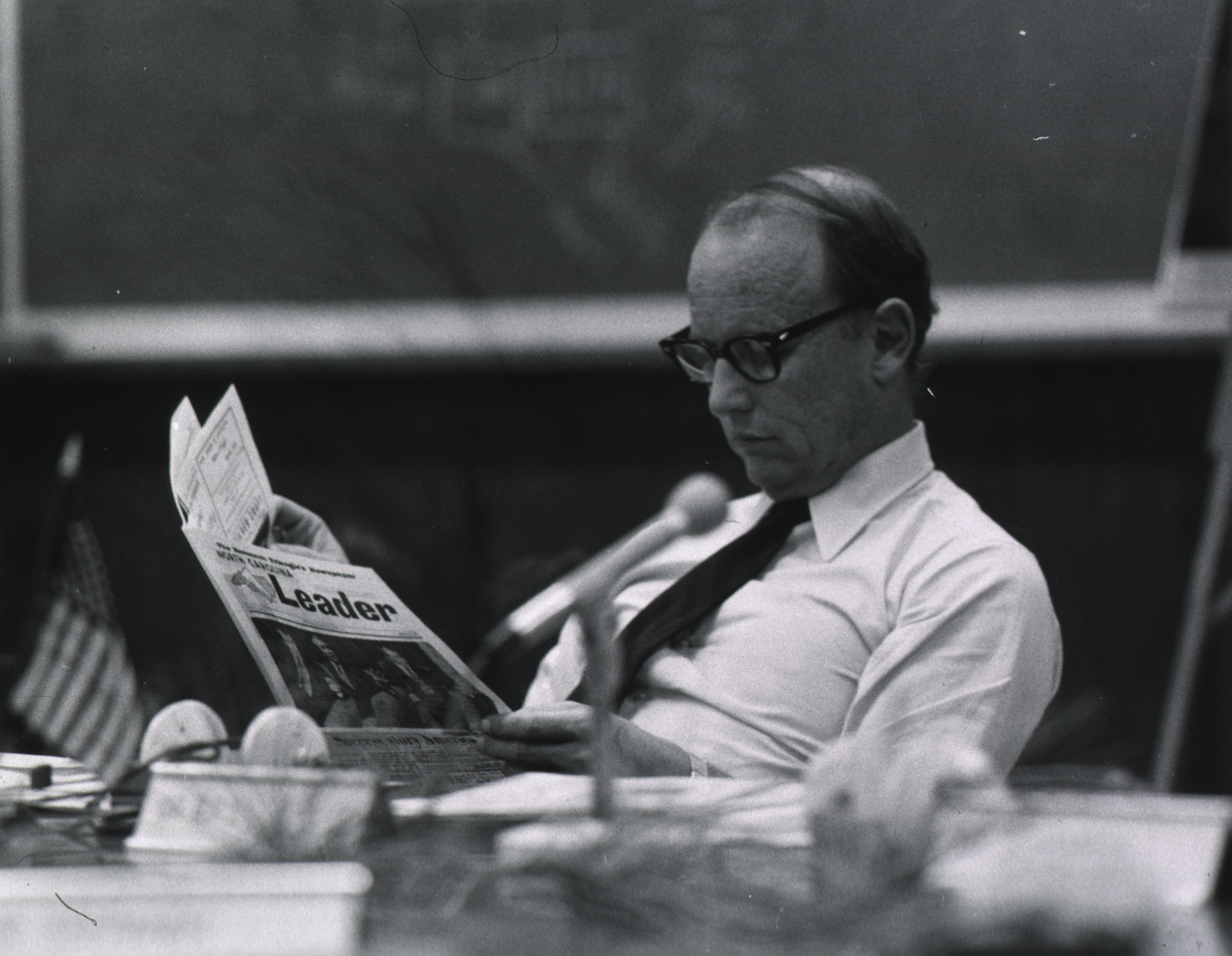 Man in glasses and tie reading a newspaper titled Leader at a desk with a microphone and an American flag.