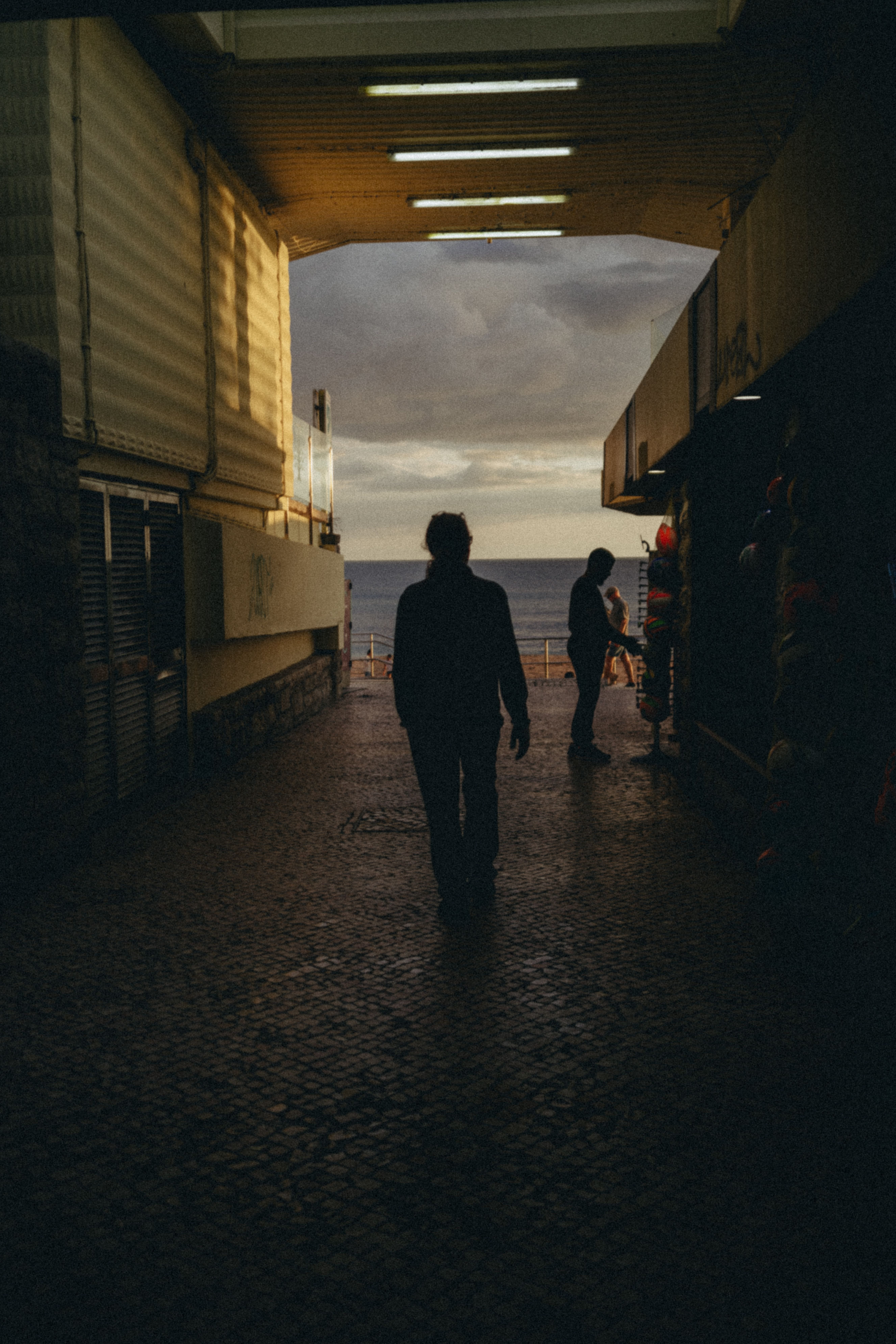 Silhouettes of two people walking in a tunnel toward a cloudy sky and ocean view at sunset.