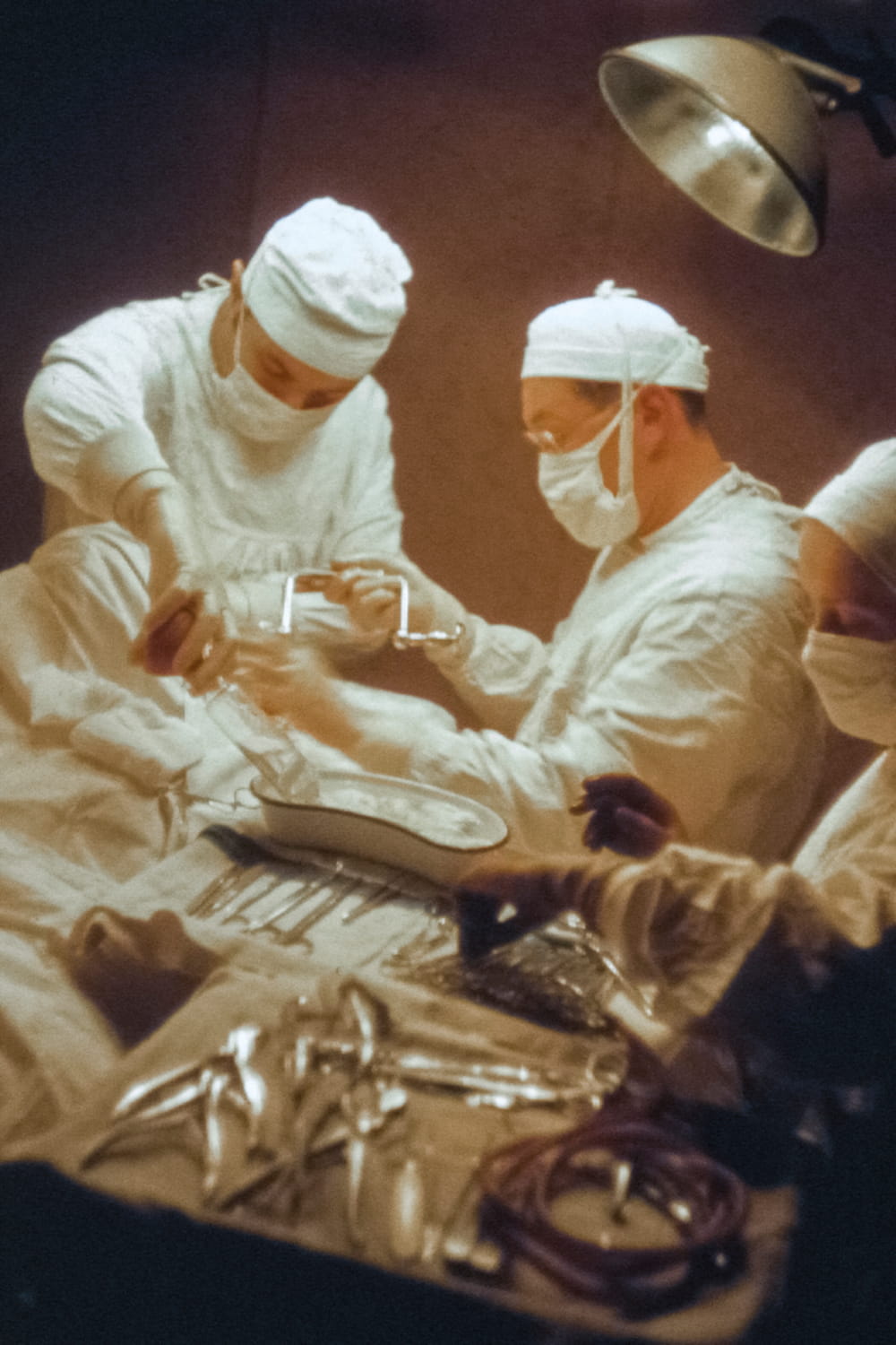 Three surgeons in white gowns and masks working with surgical tools under an overhead light.