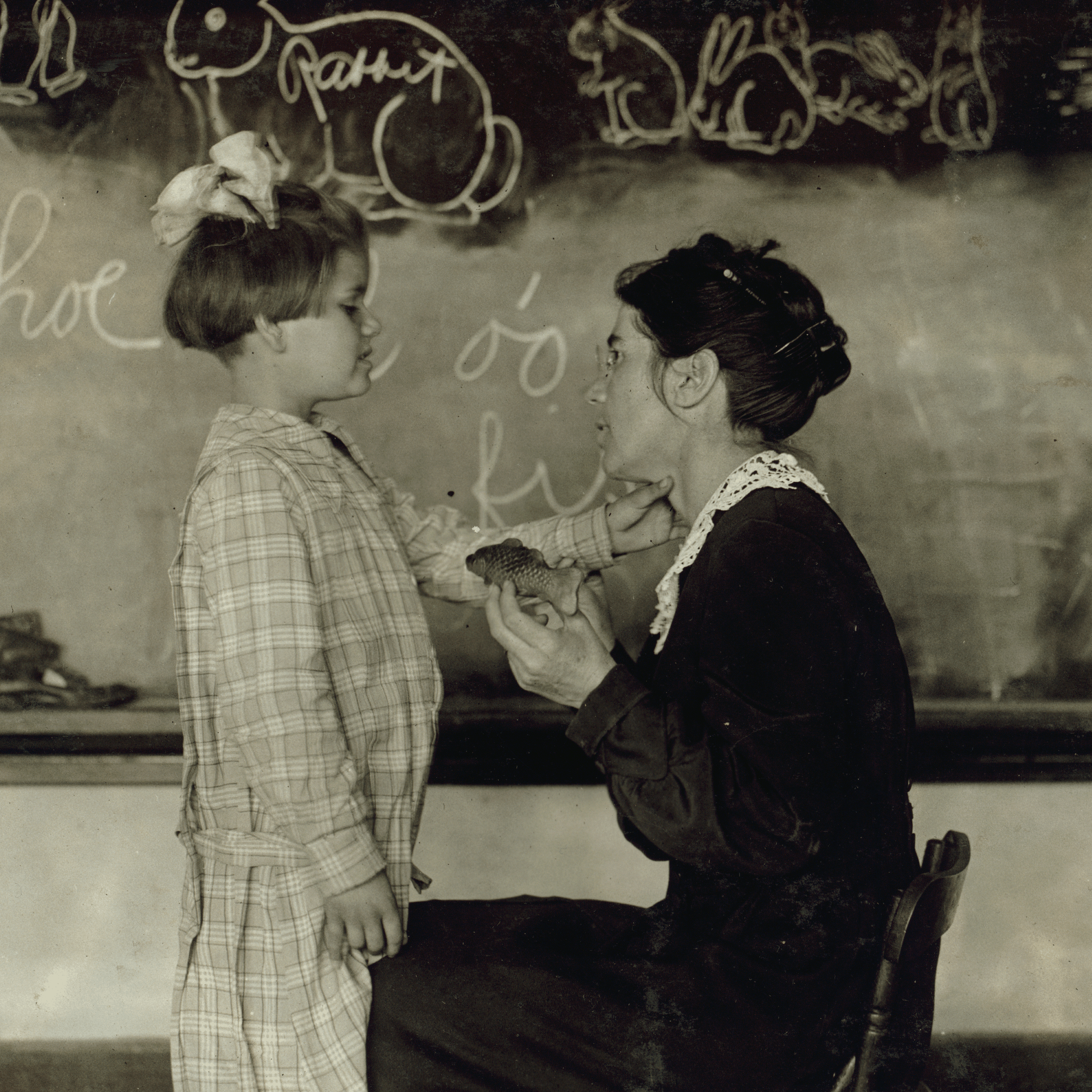 Teacher holding a fish-shaped object, engaging with a young girl in front of a chalkboard with rabbit drawings.