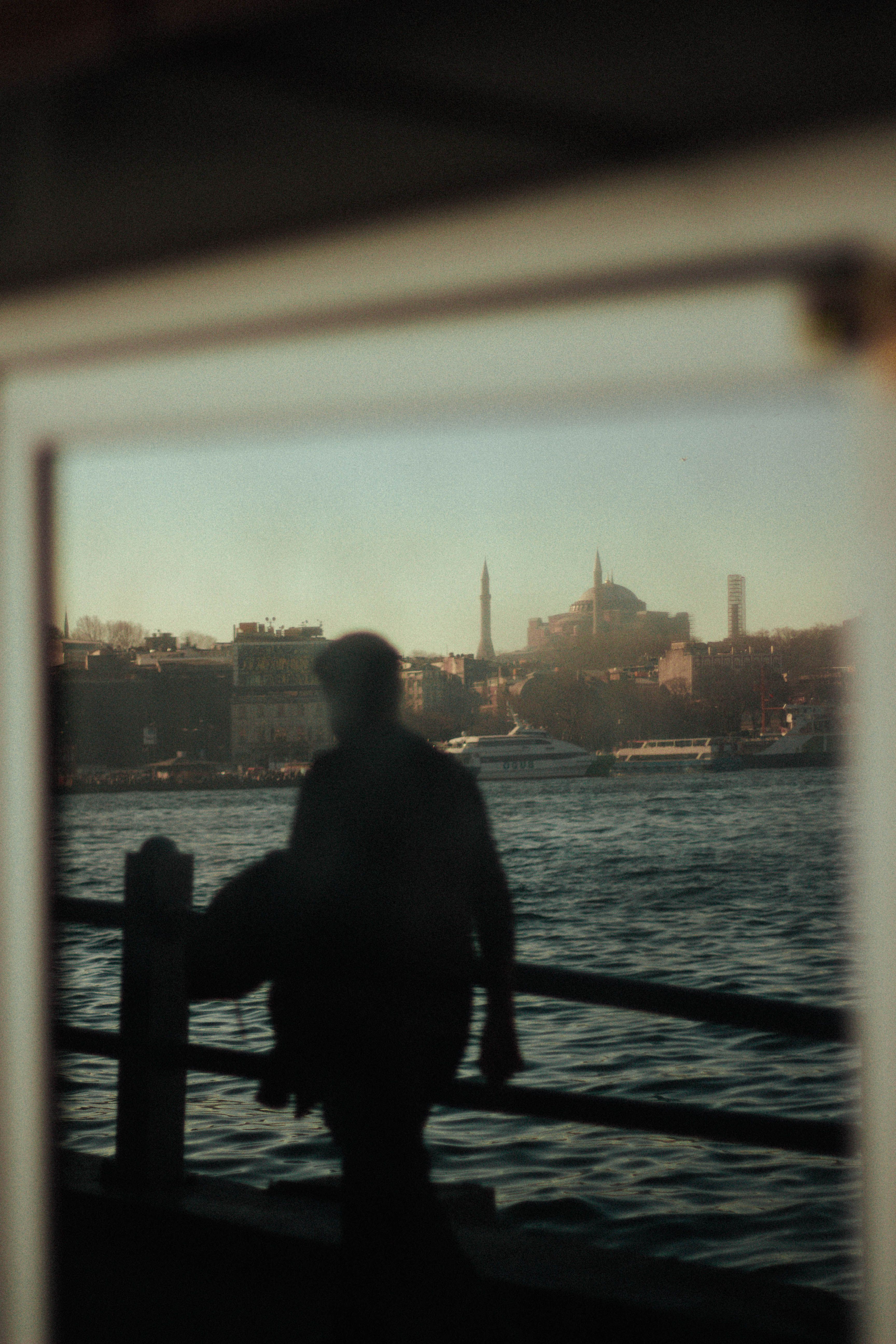 Silhouetted figure by waterfront with boats and historic mosque with minarets in background at sunset.