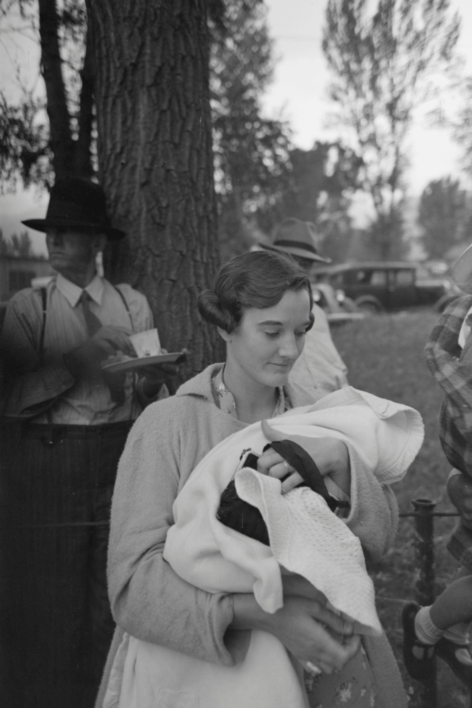 Woman holding a swaddled baby outdoors, with men and cars in the background near a tree.
