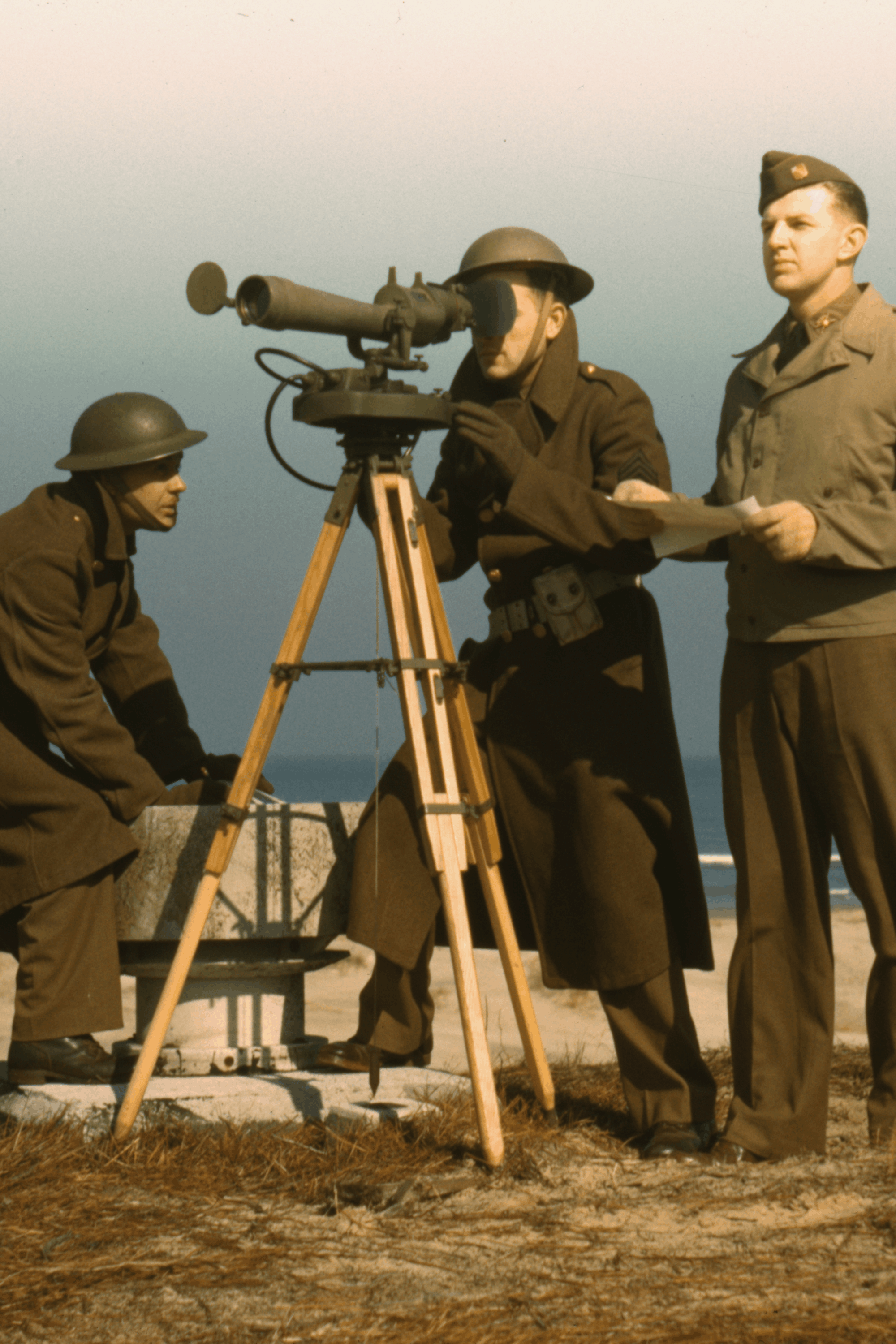 Three soldiers in World War II uniforms operating a large tripod-mounted optical rangefinder near a beach.