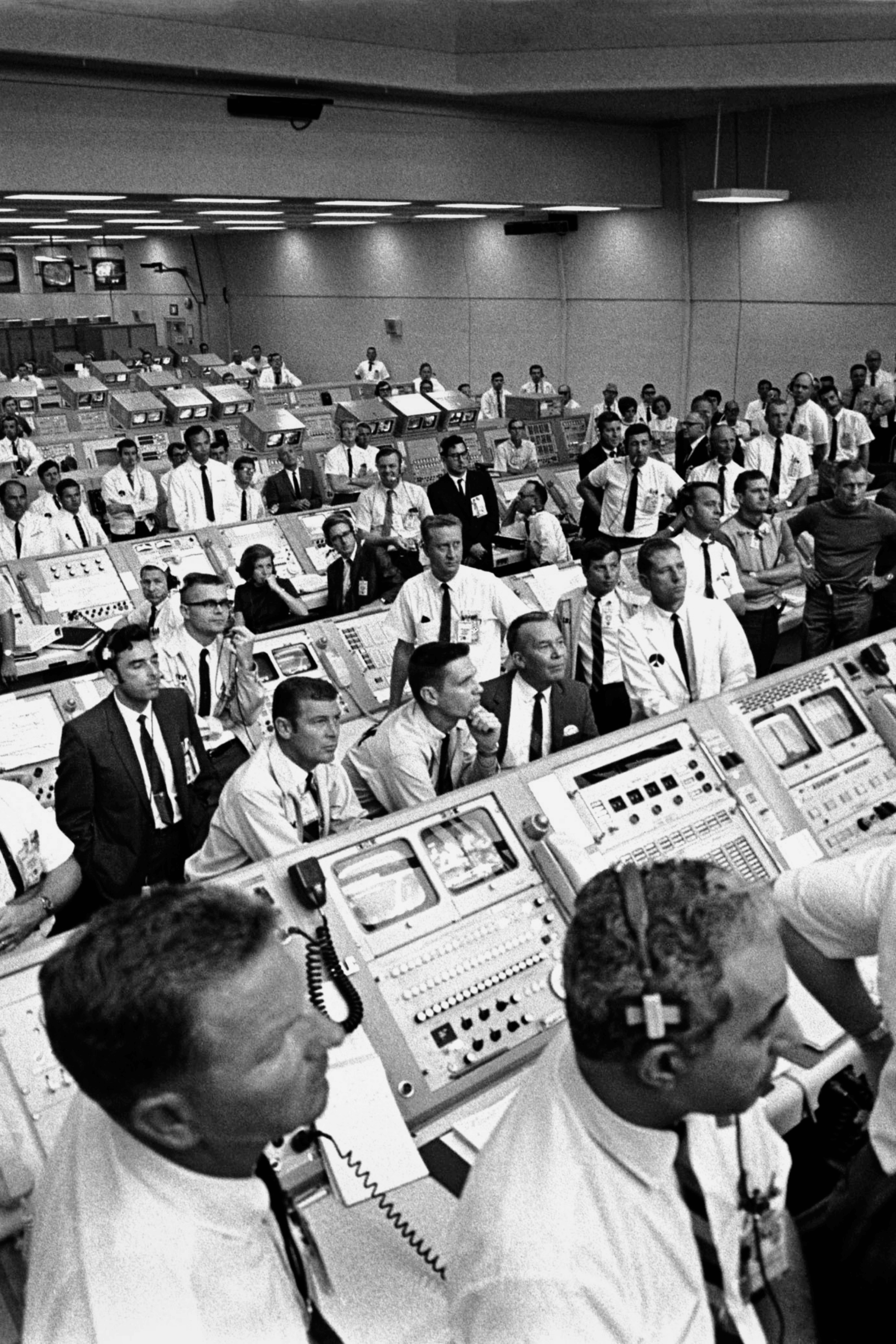 Large group of men in white shirts at control panels in a vintage mission control room.