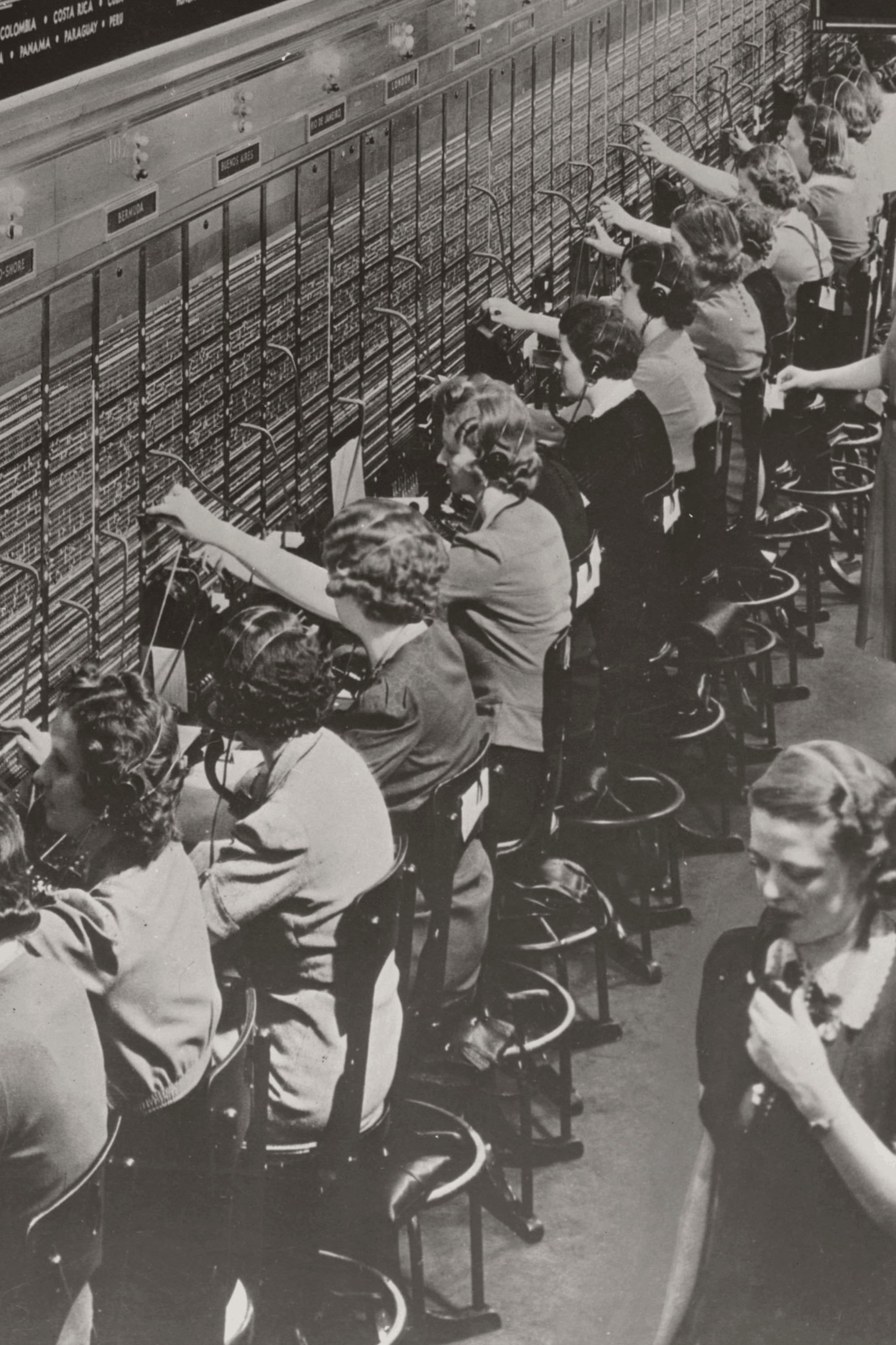 Row of vintage female telephone operators at a switchboard connecting calls with headsets and plugs.