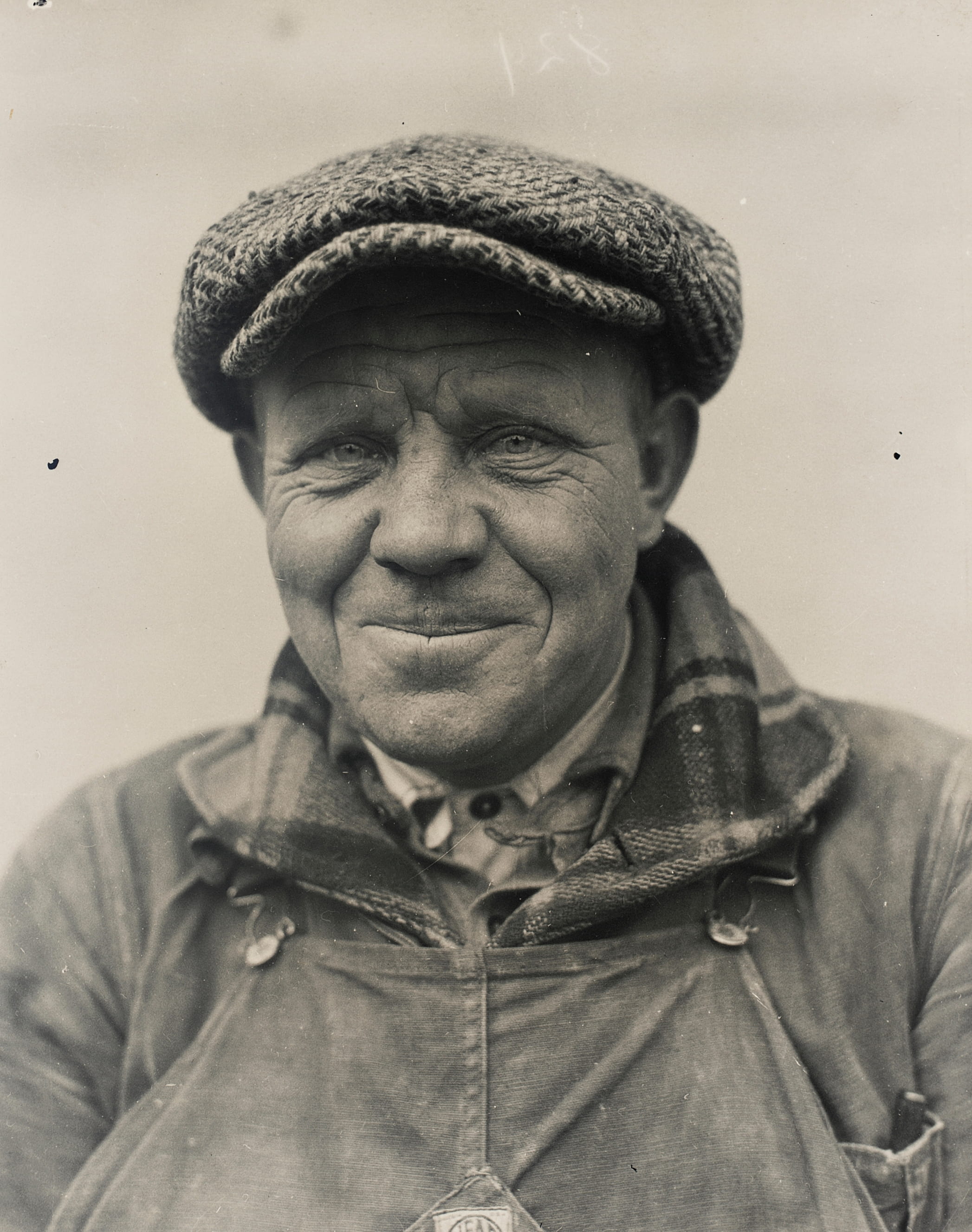 Smiling elderly man wearing a flat cap and work clothes with visible wrinkles on his face.