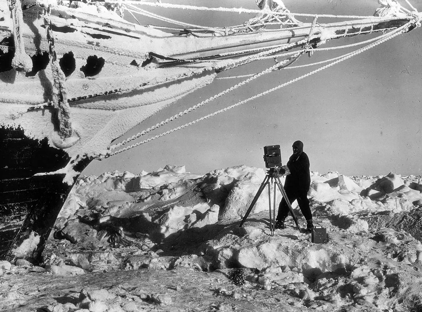 Person in cold gear photographing icy ship bow in snowy, frozen landscape with large camera on tripod.