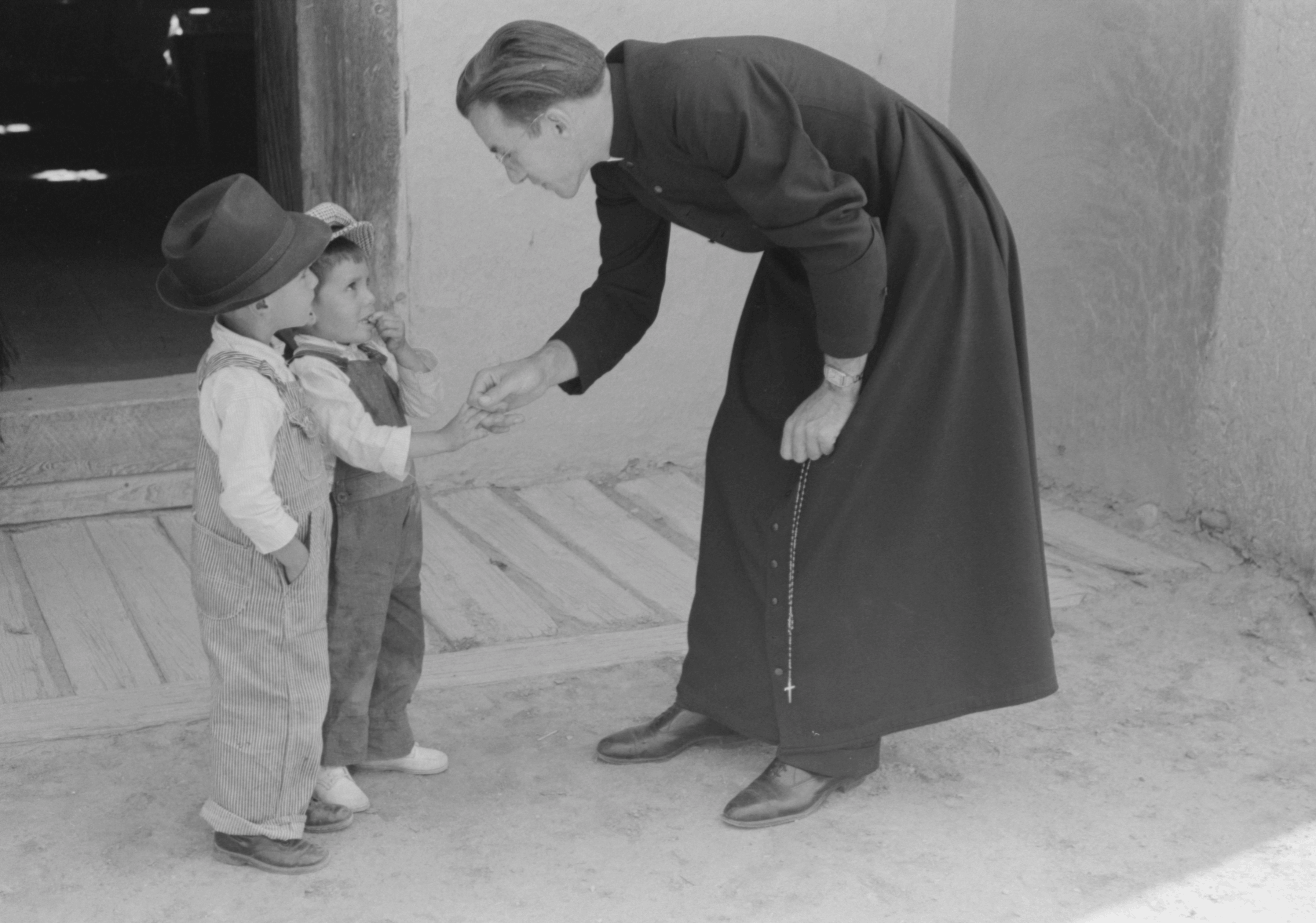 A priest bends to greet two young boys standing outside a building, holding a rosary in hand.