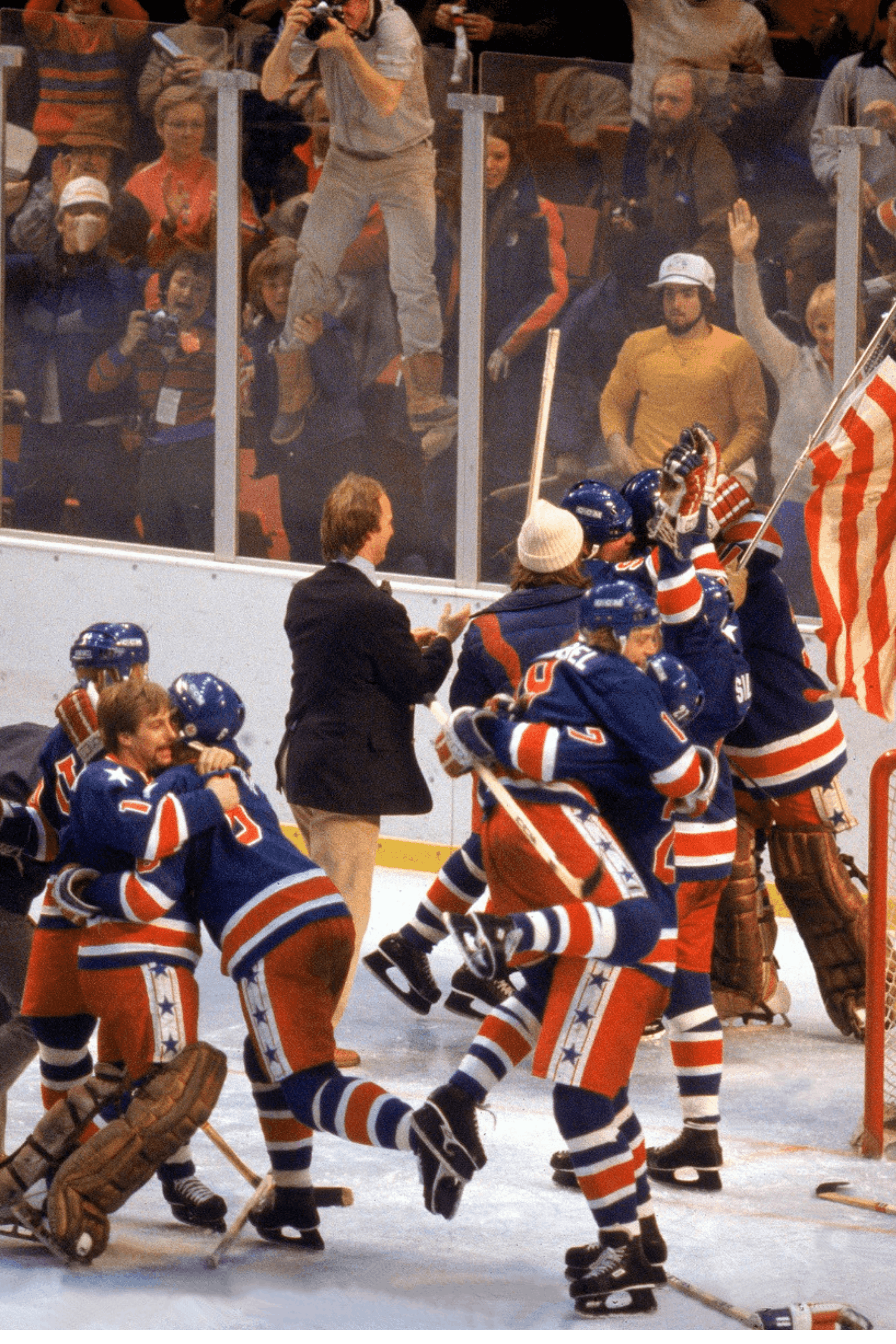 Hockey team in USA-themed uniforms celebrating a victory on the ice with fans cheering behind glass.