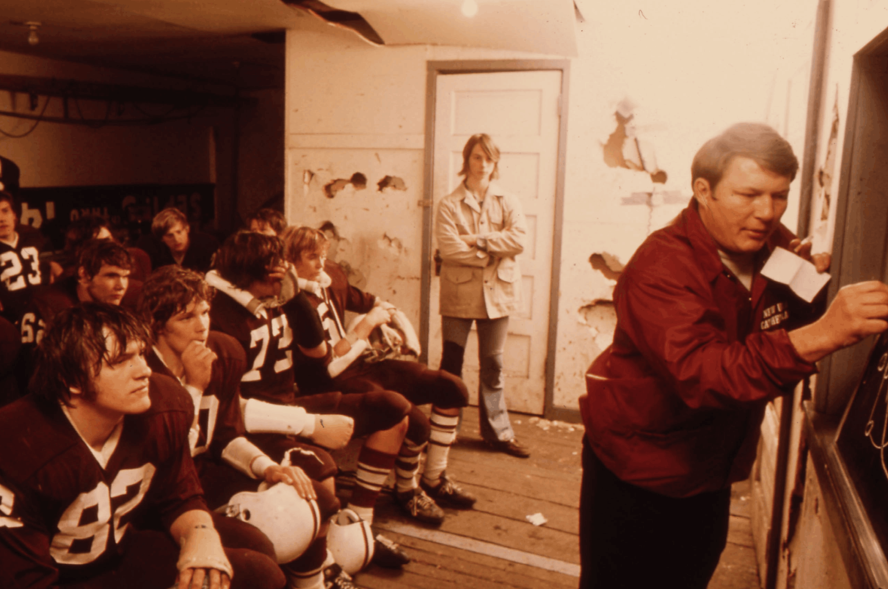 Football coach drawing a play on a chalkboard while players in uniforms watch in locker room.