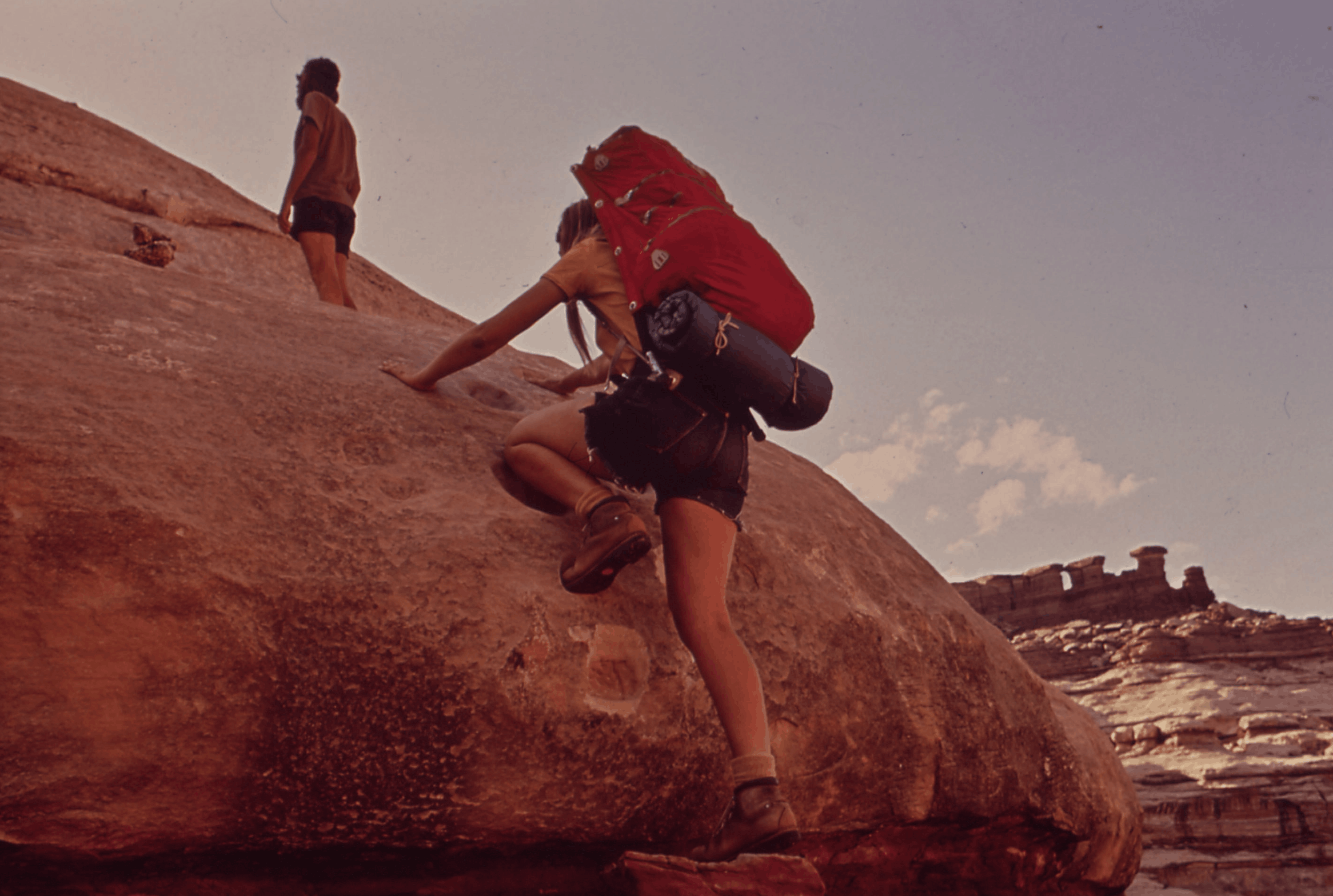 Two hikers climbing a large reddish rock formation under a clear sky.