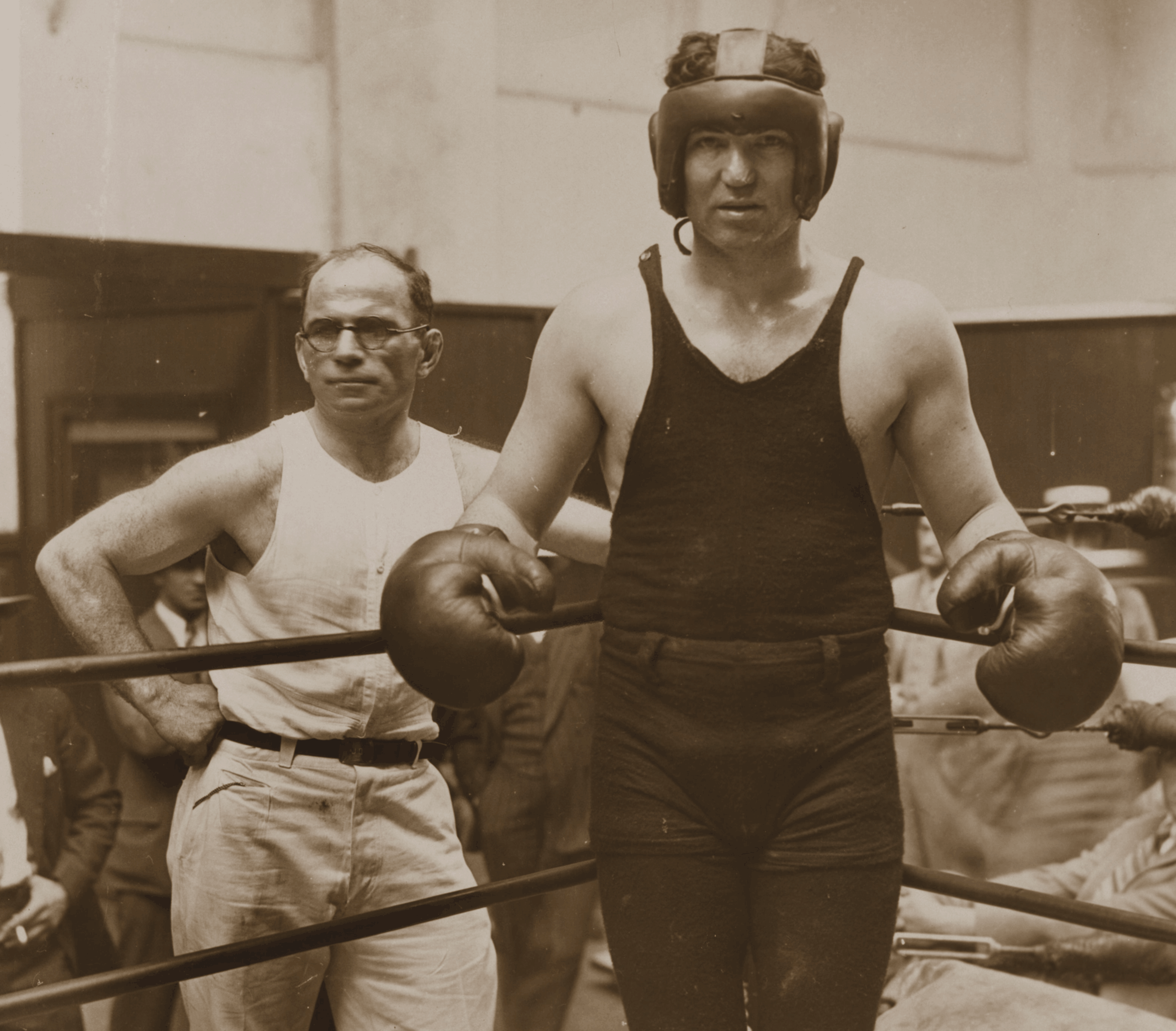 Vintage photo of a boxer in gear and gloves standing in ring beside a man in a sleeveless white shirt.