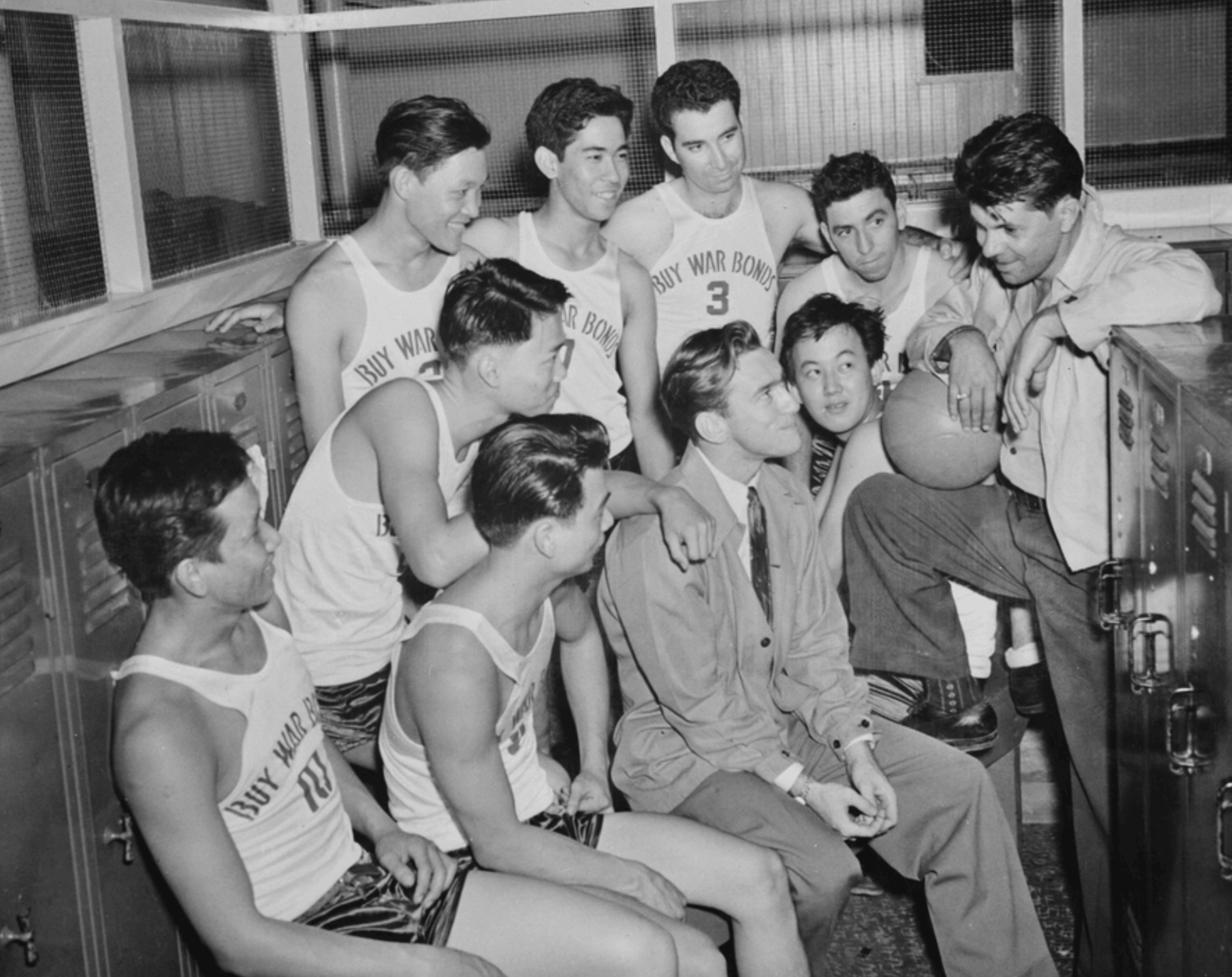 Black and white photo of a basketball team in 'Buy War Bonds' jerseys gathered in a locker room talking.