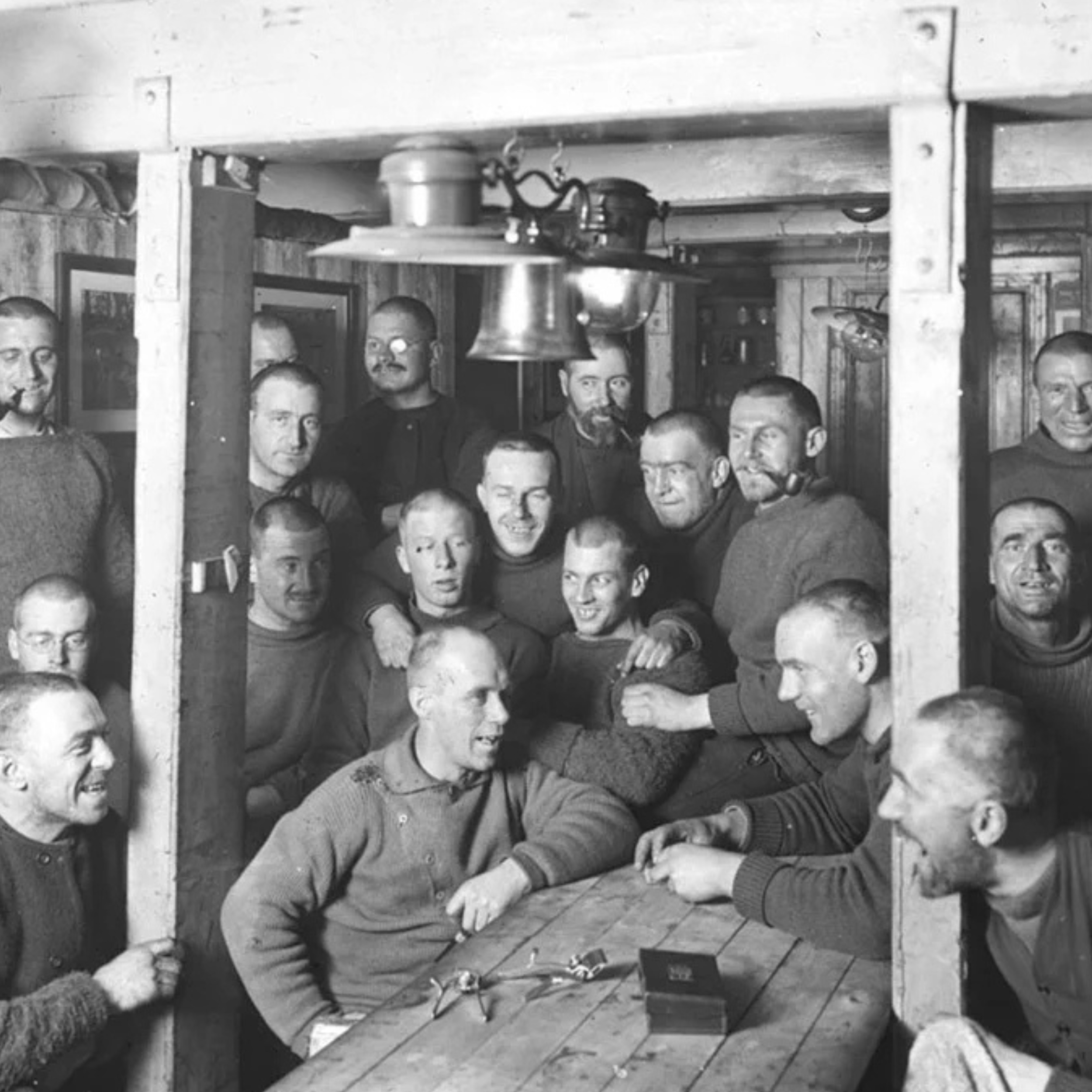 Group of men in sweaters gathered around a wooden table inside a rustic room, some smoking pipes.