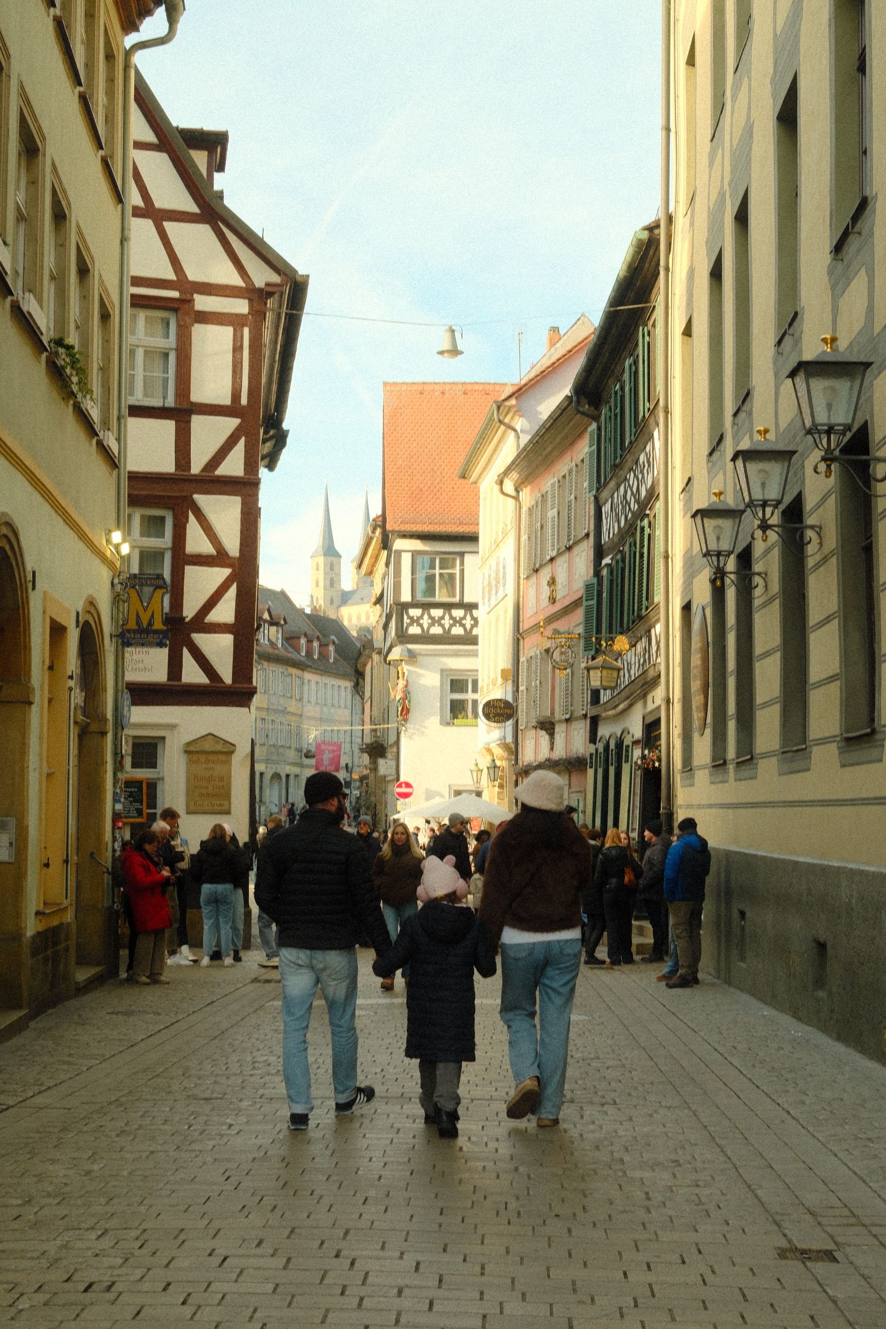 A family of three holding hands walking down a cobblestone street lined with historic buildings.
